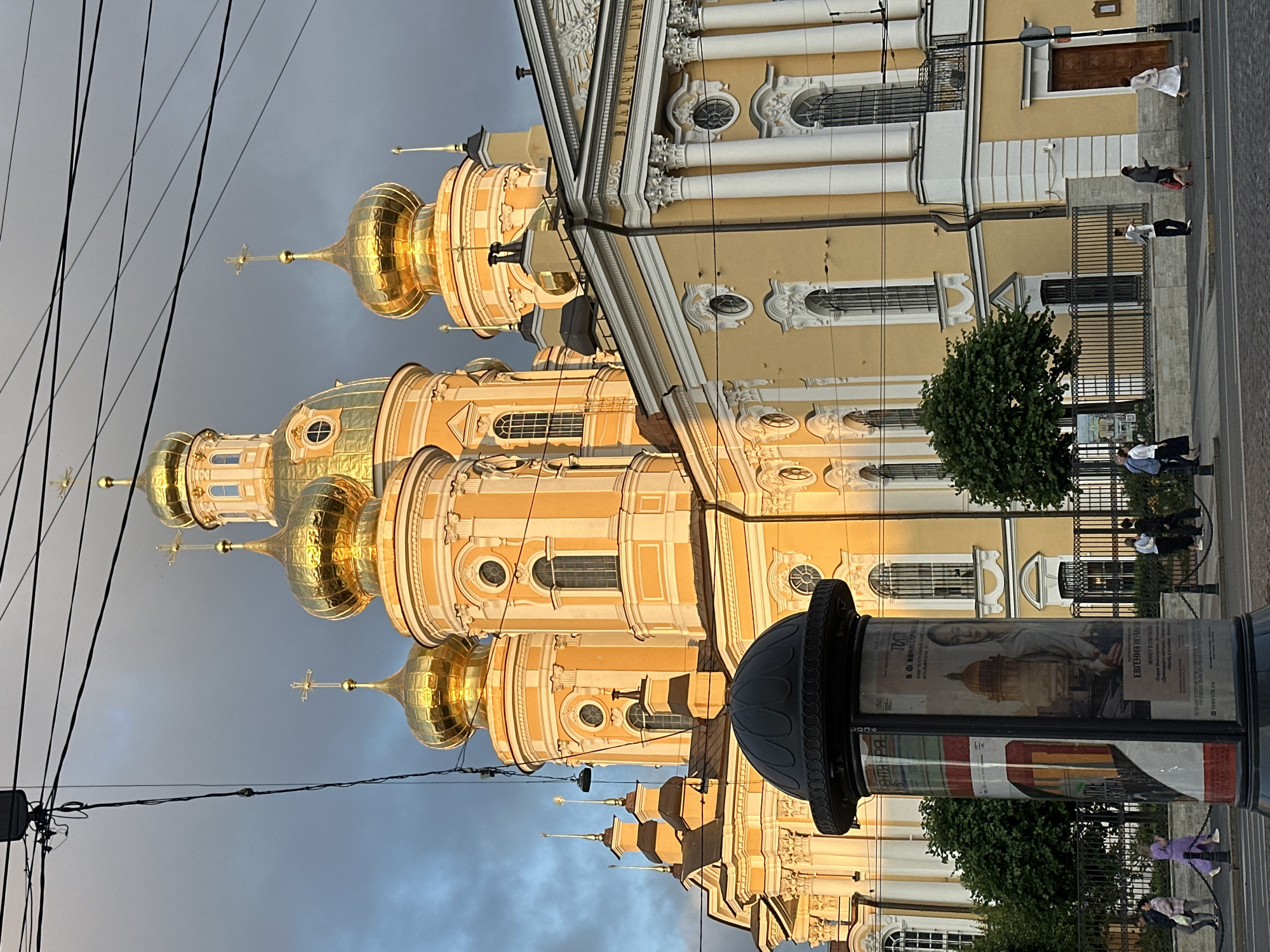 The image depicts a bright yellow church with multiple golden domes against a cloudy sky in Saint Petersburg, Russia. In the foreground, a dark cylindrical advertising pillar stands on a cobblestone street, with several people walking on the sidewalk. The church features ornate architecture, including columns and arched windows, and appears to be illuminated by the late afternoon sun. Power lines are visible across the top of the frame, and trees line the sidewalk behind a black metal fence.
