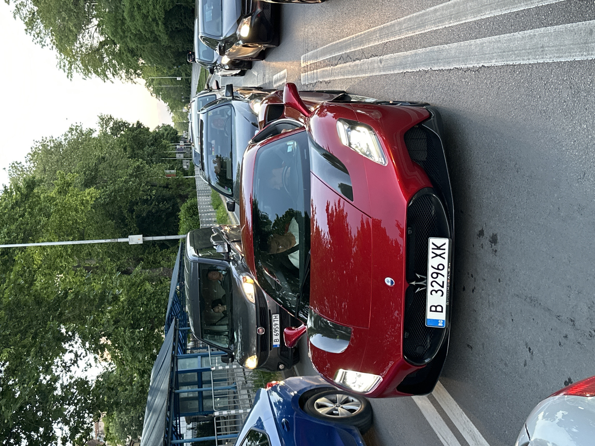 The image shows a street scene in Bulgaria with several cars in a traffic jam. The closest car is a red Maserati with a Bulgarian license plate "B 3296 XK". Behind the Maserati is a dark-colored Fiat Doblo with license plate "B 6959 TH". Further back are several other cars, and the street is lined with trees. On the left side of the street is a bus stop. The image does not provide any direct indication that this is in Varna.