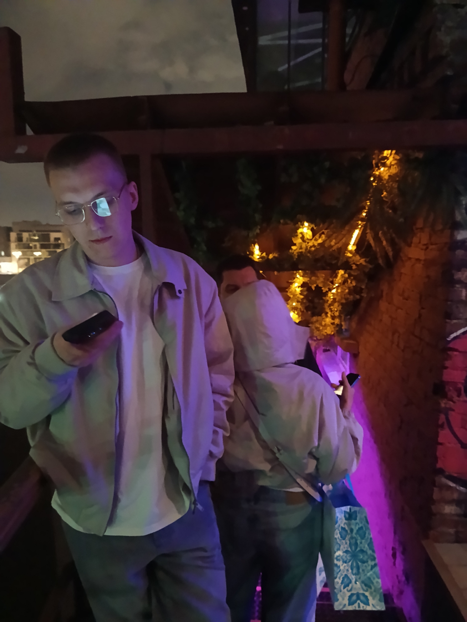 A man with short hair and glasses looks at a smartphone in his hand. He wears a white T-shirt and a light jacket. Behind him are two figures, one wearing a light jacket with the hood up and holding a smartphone and a shopping bag. The location appears to be an outdoor area near a brick wall with graffiti. Some foliage with string lights is visible. A wooden structure is above the people. The city is Moscow, Russia.