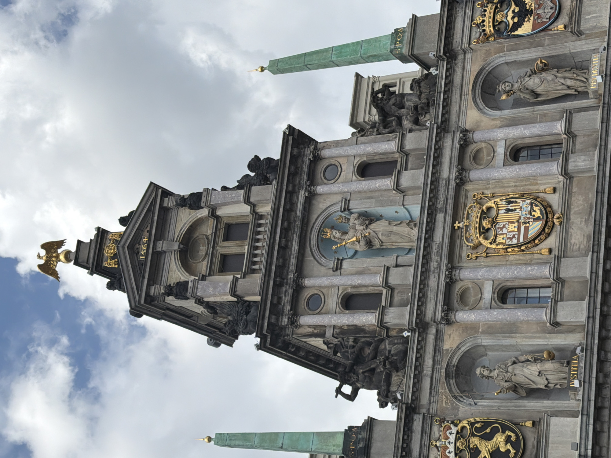 This is a low-angle, medium shot of the Antwerp City Hall, located in Antwerp, Belgium. The historic building features a prominent clock tower topped with a golden eagle statue. Niches in the facade hold statues of figures, including what appears to be a representation of Justice, identified by the inscription "IVSTITIA" on its pedestal. The building's architecture is ornate, with columns, intricate carvings, and coat of arms crests. Two green obelisks flank the structure, one on the left and one on the right, each topped with a golden finial. The sky above is partly cloudy.