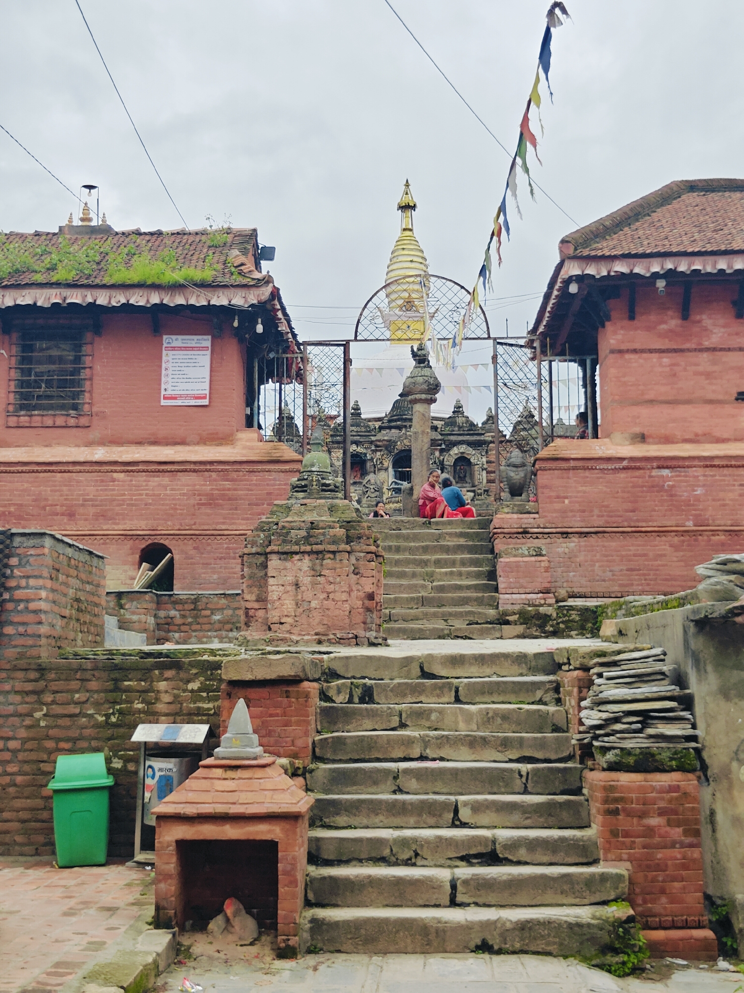 The image depicts a view of the entrance to a temple complex in Kirtipur, Nepal. In the foreground, a wide, stone staircase leads upwards. To the left of the stairs is a green trash can and a small, brick structure with a pointed roof, beneath which a person is seated. Further up the stairs, a metal gate leads into the temple complex. Beyond the gate, prayer flags are strung across the sky. A golden stupa with a conical spire is visible behind the gate, topped with a finial. Two brick buildings with tiled roofs flank the central stupa, with lush green vegetation growing on the roof of the left building. Several people are seated on the steps and near the gate. The overall atmosphere suggests a spiritual and cultural site.