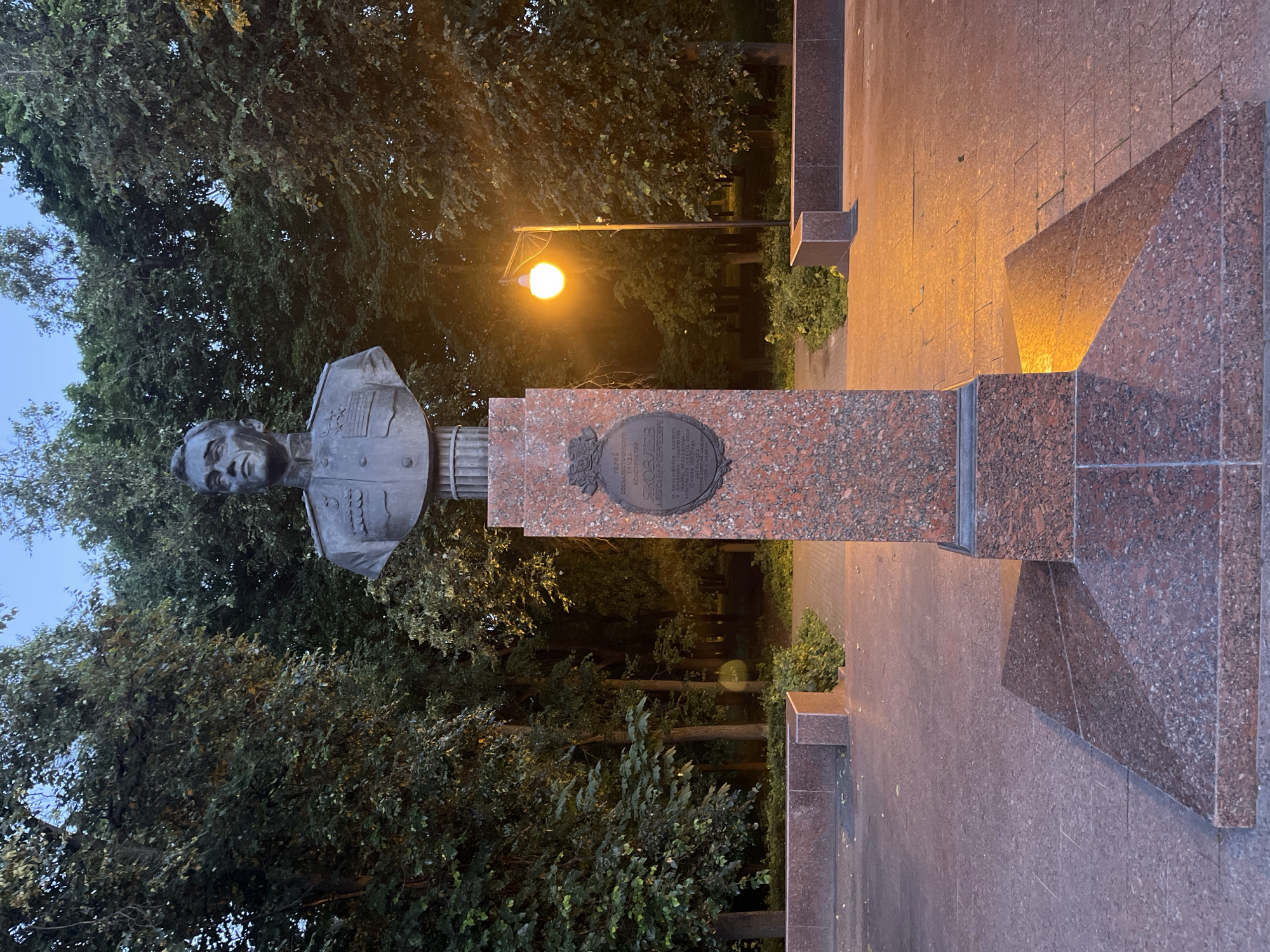 A bust of a man in a military uniform is centered on a granite pedestal in a park. The bust is made of dark metal and depicts a man with short, dark hair, looking to his left. He wears a jacket with medals and epaulets. The pedestal is made of red granite with a plaque on its front. A lamppost with a globe-shaped light casts a warm glow on the scene. The background consists of trees and a clear sky. The ground around the monument is paved with red granite tiles, and there are low granite benches. The overall scene suggests a public memorial or monument in a park setting. The city is Unknown, Unknown.