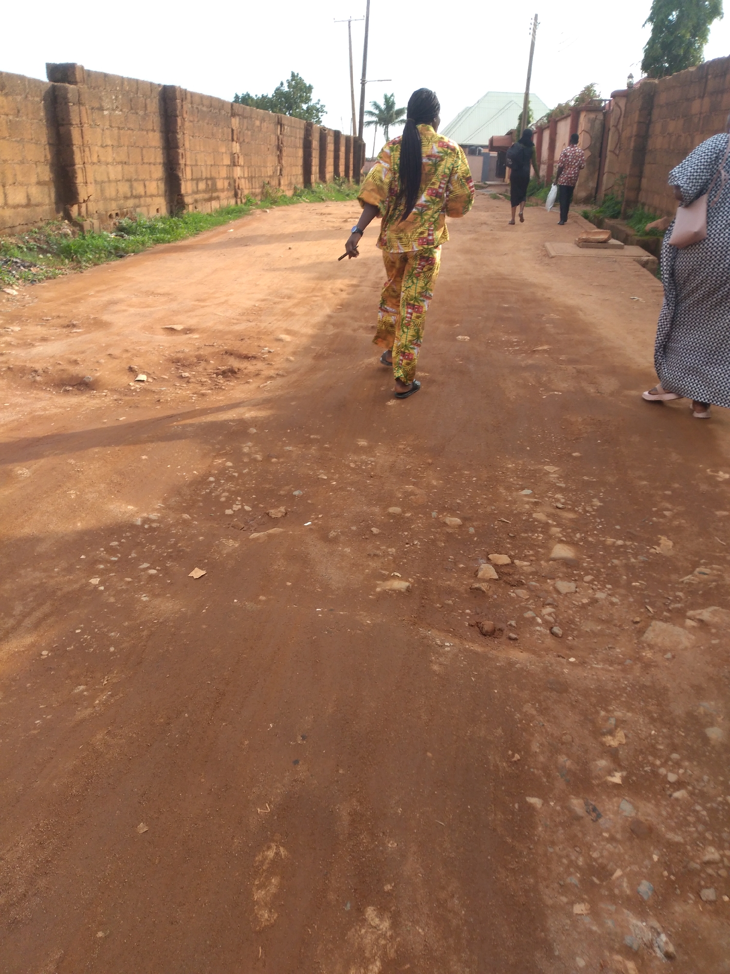 A group of four individuals are walking along a dirt road in Jos, Nigeria. In the foreground, a person wearing a yellow and green patterned outfit with a ponytail walks away from the camera. To the right, another person in a black and white checkered dress is visible. Further down the road, two more individuals are walking away; one in a black dress and the other in a patterned shirt and dark pants. The road is unpaved and reddish-brown, with scattered stones and debris. On the left side of the road, there is a tall, weathered brick wall. On the right, there are more walls and buildings in the background. The sky is overcast.