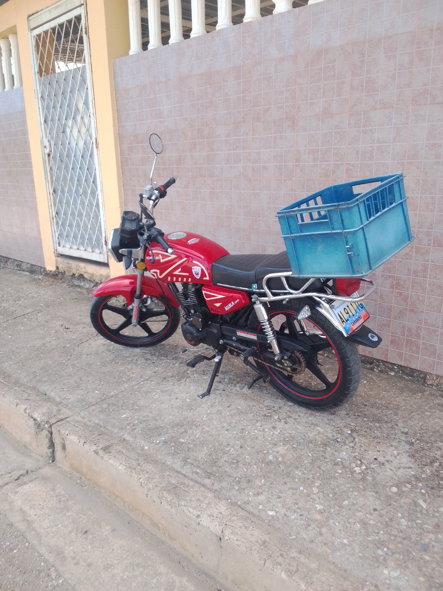 A red motorcycle with a blue plastic crate attached to the back is parked on a concrete sidewalk next to a light tan building with a white gate. The motorcycle has a license plate that reads "AL9111G". The building has a tiled wall in a light pinkish-tan color. The sidewalk is made of concrete slabs and has a curb. The scene is outdoors in daylight.