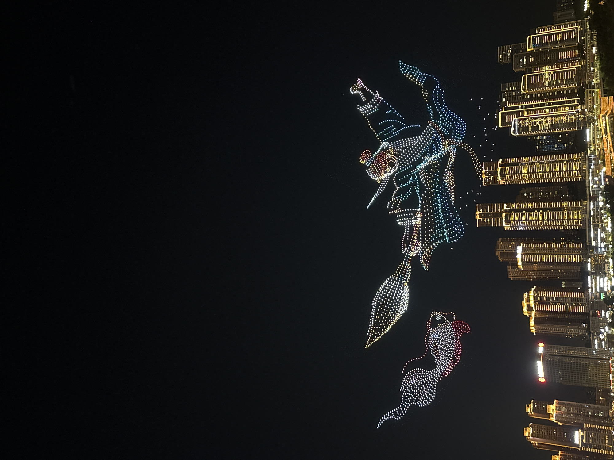 An aerial drone light show takes place over the Yuzhong District of Chongqing, China, on a dark night. The sky is filled with illuminated drone formations, depicting a stylized dragon and a fish-like creature. The dragon, predominantly white and blue with accents of red and yellow, is positioned in the upper right, appearing to fly forward. To its left, a red and white fish-like formation, with fins and a tail, is depicted. Below the formations, the Yuzhong District skyline is illuminated with numerous tall buildings featuring yellow and white lights, creating a vibrant urban panorama. The light show is in progress above the city.