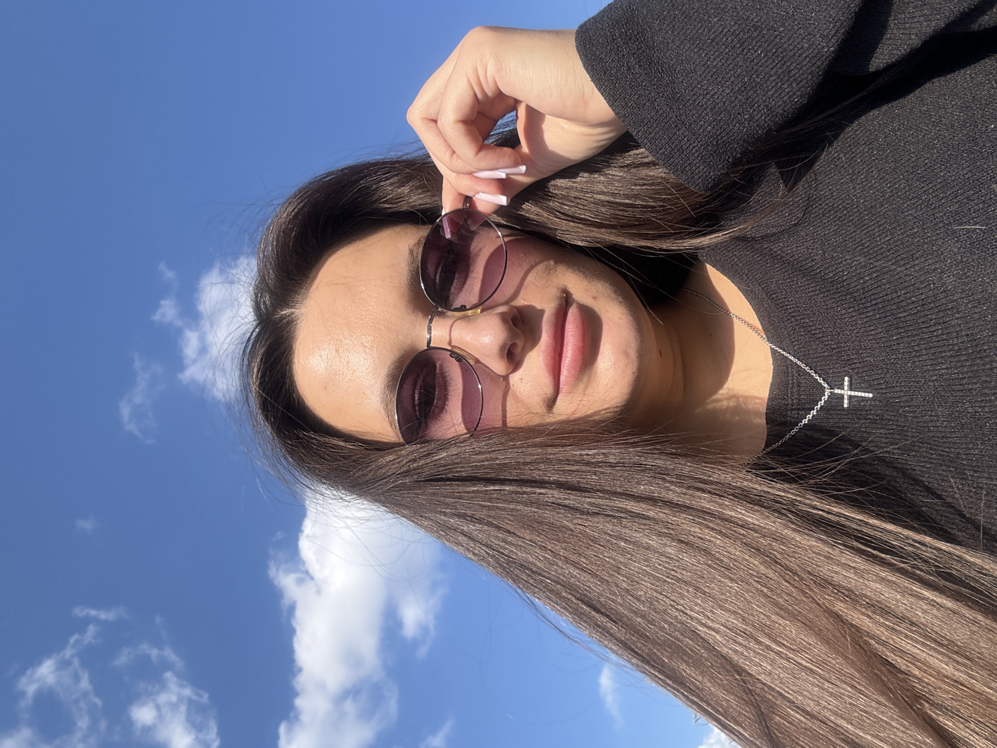 A young woman with long brown hair is holding purple-tinted round sunglasses in her right hand. She is wearing a black long-sleeved shirt and a silver cross necklace. The background shows a blue sky with white clouds. The location is likely Akulovo (Gp Kubinka), Russia, as requested, but this is inferred and not directly verifiable from the image. The focus is on the woman and her pose with the sunglasses.