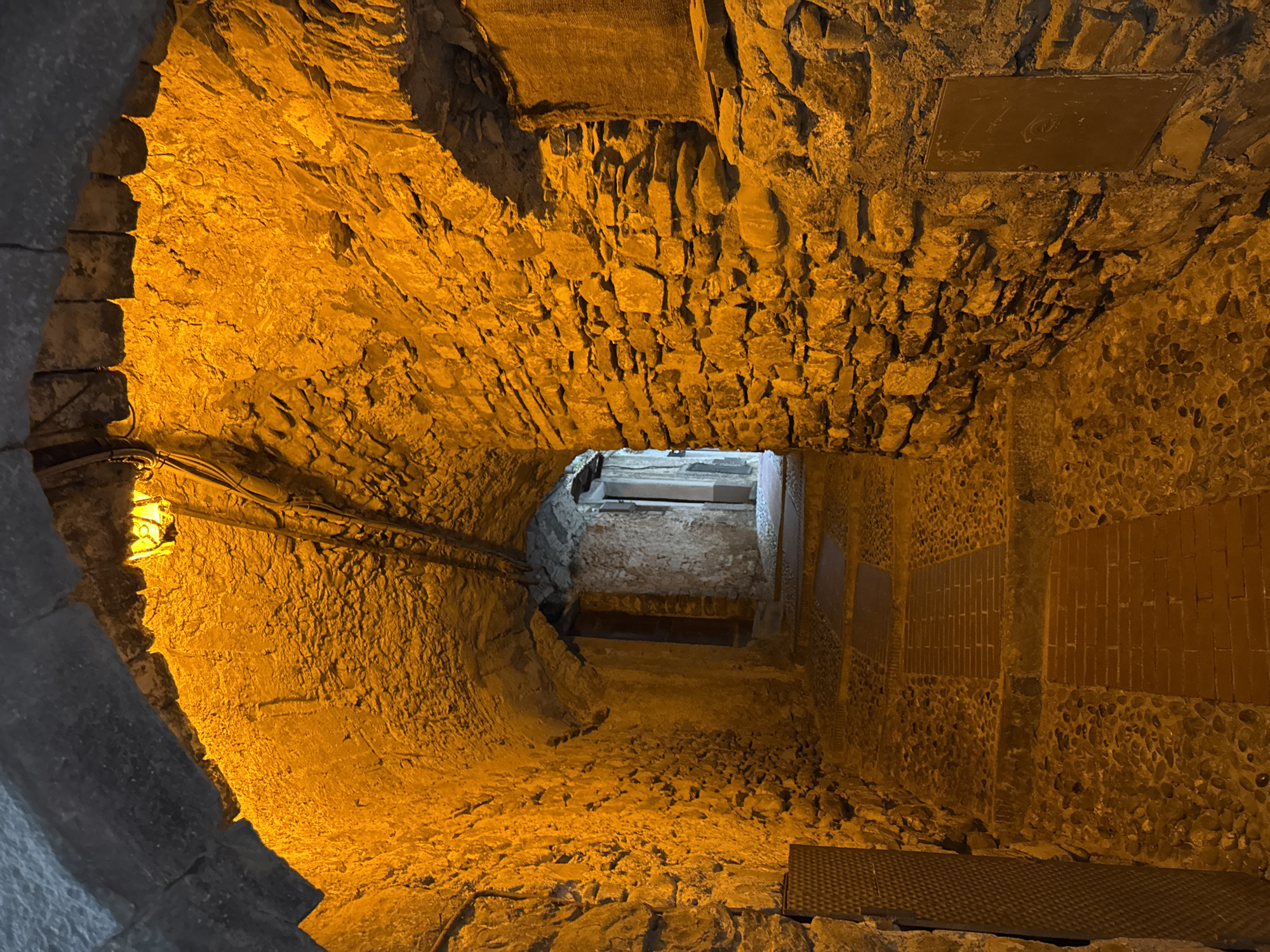 This is a nighttime, medium-shot, eye-level view of an ancient stone passageway in Dolceacqua, Italy. The arched tunnel is illuminated by a warm, orange light emanating from a single fixture at the apex of the tunnel's ceiling, casting long shadows and highlighting the rough texture of the stone walls. The passageway slopes gently upwards and curves slightly to the right, leading the eye towards a distant, dimly lit opening. The floor is paved with a mixture of cobblestones and brickwork, forming a stepped pathway. The walls are constructed of irregular, stacked stones, some of which appear to be weathered and ancient. A dark, metallic grate is visible on the left side of the passageway, near the foreground. Further down the tunnel, wooden doors are set into the stone walls. The overall impression is one of historical depth and atmospheric mystery.