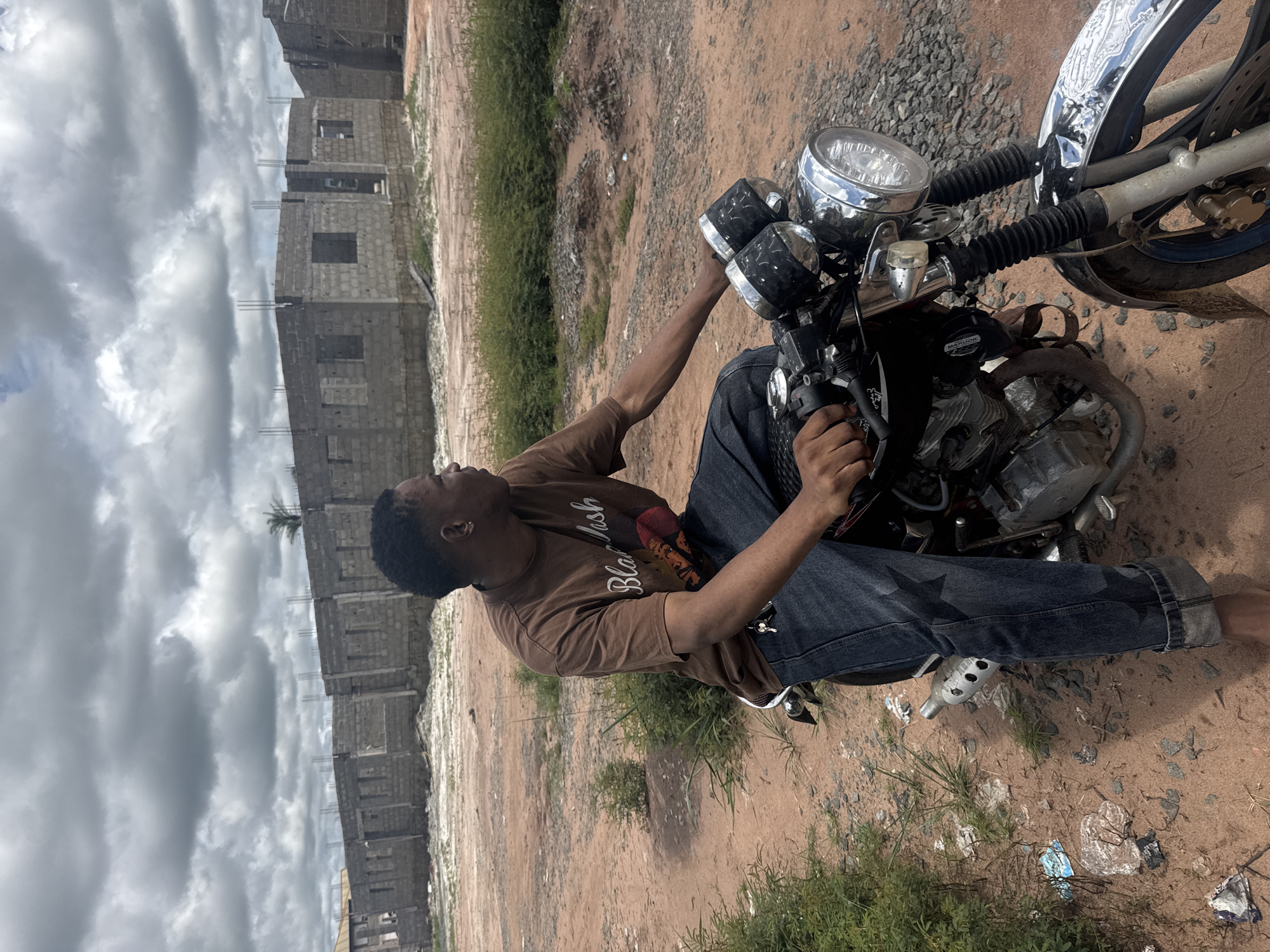 A young Black male, wearing a brown t-shirt with "Black Nash" printed on it and dark wash jeans with a star pattern, is sitting on a motorcycle. He is looking to his right, away from the camera. The motorcycle is dark-colored with chrome accents, including the handlebars and dual headlights. The scene appears to be an outdoor, undeveloped area in Ihiagwa, Nigeria, characterized by a dusty, reddish-brown ground and sparse green vegetation. In the background, there are several unfinished brick buildings, suggesting a construction site. The sky is overcast with dramatic, dark clouds. There is some scattered debris on the ground, including plastic bags and wrappers.