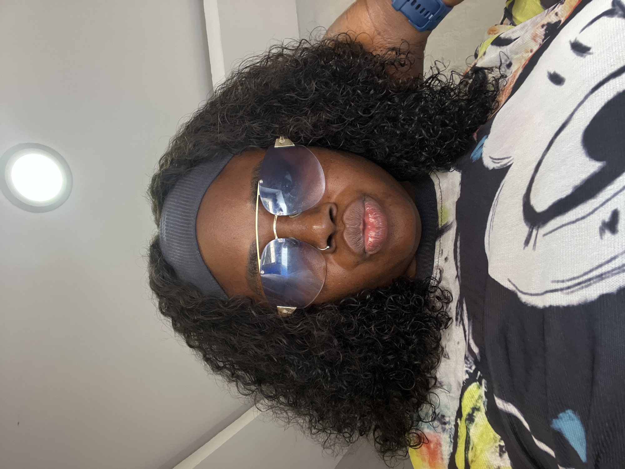 A medium shot shows a dark-skinned person wearing a gray headband, large aviator sunglasses with blue lenses, and a black t-shirt with a colorful abstract pattern of Mickey Mouse. The person has curly black hair and is puckering their lips, with a nose ring visible. They are touching their head with their right hand. A blue digital watch is on their left wrist. The background appears to be an indoor setting with a white ceiling and a circular light fixture. There is no indication of the specific location within Abuja, Nigeria.