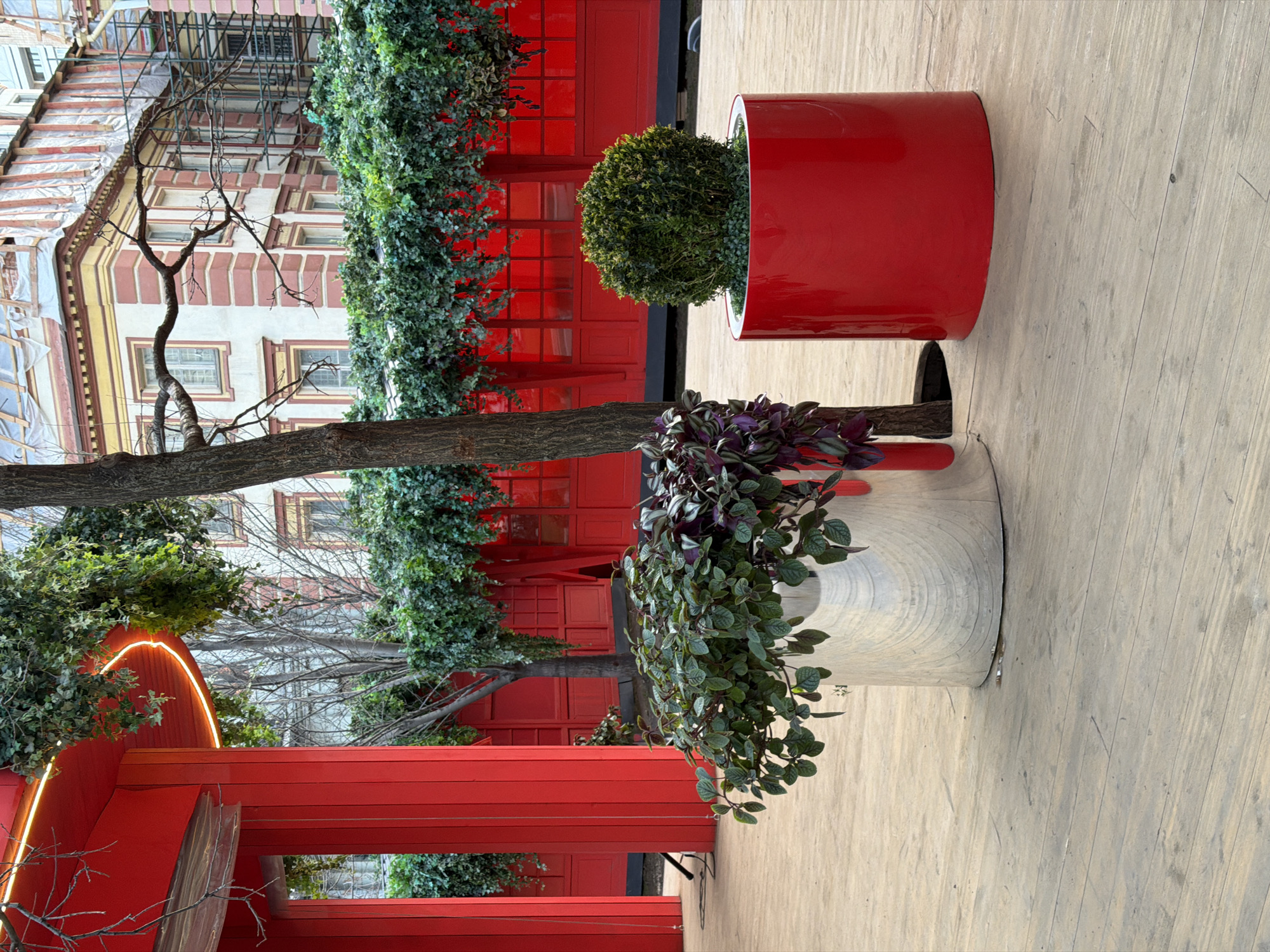 The image depicts an outdoor setting in Moscow, Russia, featuring decorative elements and architectural details. In the foreground, there are two cylindrical planters: one red and one silver. The red planter contains a round, green shrub, while the silver planter holds a leafy plant with a mix of green and purple foliage. A tree trunk is visible, seemingly running through both planters.
In the background, there is a red architectural structure with ivy climbing on it. Further back, a building with white and brown facade details is visible, along with some bare trees. The ground is covered with light-colored wooden planks. There is no indication of people in the image.