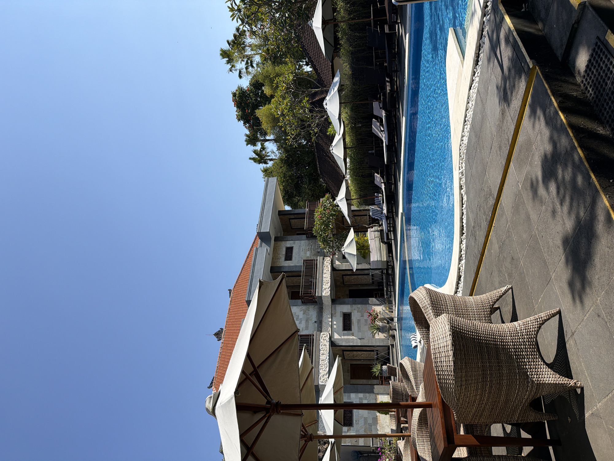 This image captures a serene resort scene in Denpasar, Indonesia, under a clear blue sky. In the foreground, several wicker chairs surround a wooden table, casting shadows on the dark tiled patio. Beyond this seating area, a bright blue swimming pool dominates the mid-ground, its surface disturbed by gentle ripples. Along the pool's edge, white pebbles are interspersed with light-colored paving stones. Several white umbrellas provide shade over lounge chairs arranged on the opposite side of the pool.
A multi-story building, constructed with stone and featuring traditional Balinese architectural elements like ornate carvings and terracotta roof tiles, forms the backdrop. Balconies with wooden railings are visible on the upper floors. Lush green trees and palm trees add to the tropical ambiance. The overall impression is one of a tranquil vacation setting, inviting relaxation and leisure. There are no people visible in the image.
