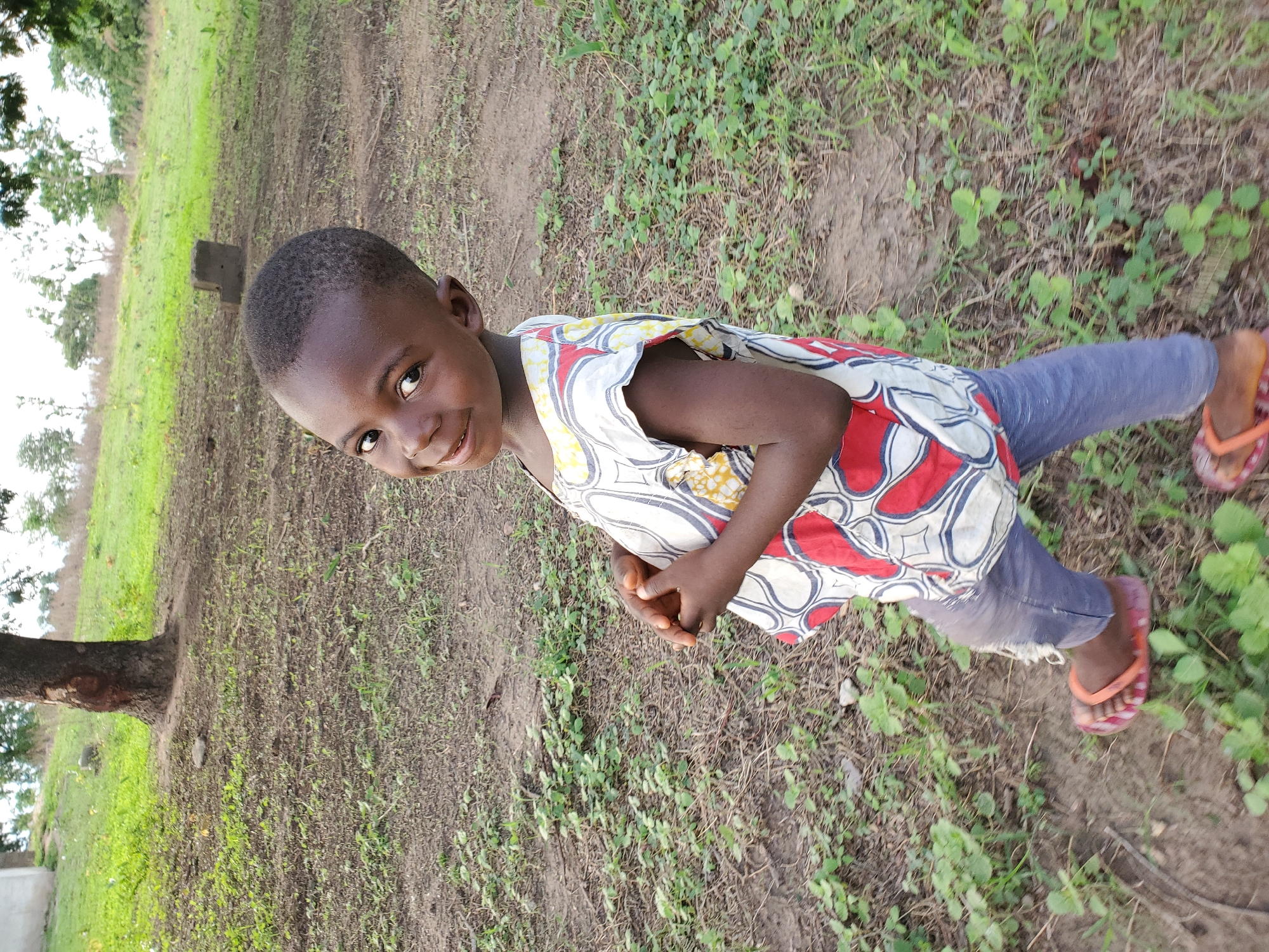 A young child stands outdoors in Mararaba, Nigeria. The child has short, dark hair and wears a light-colored sleeveless top with red, white, and yellow patterns. They also wear blue jeans and orange sandals. The environment is mostly grass and dirt, with some trees visible in the background.