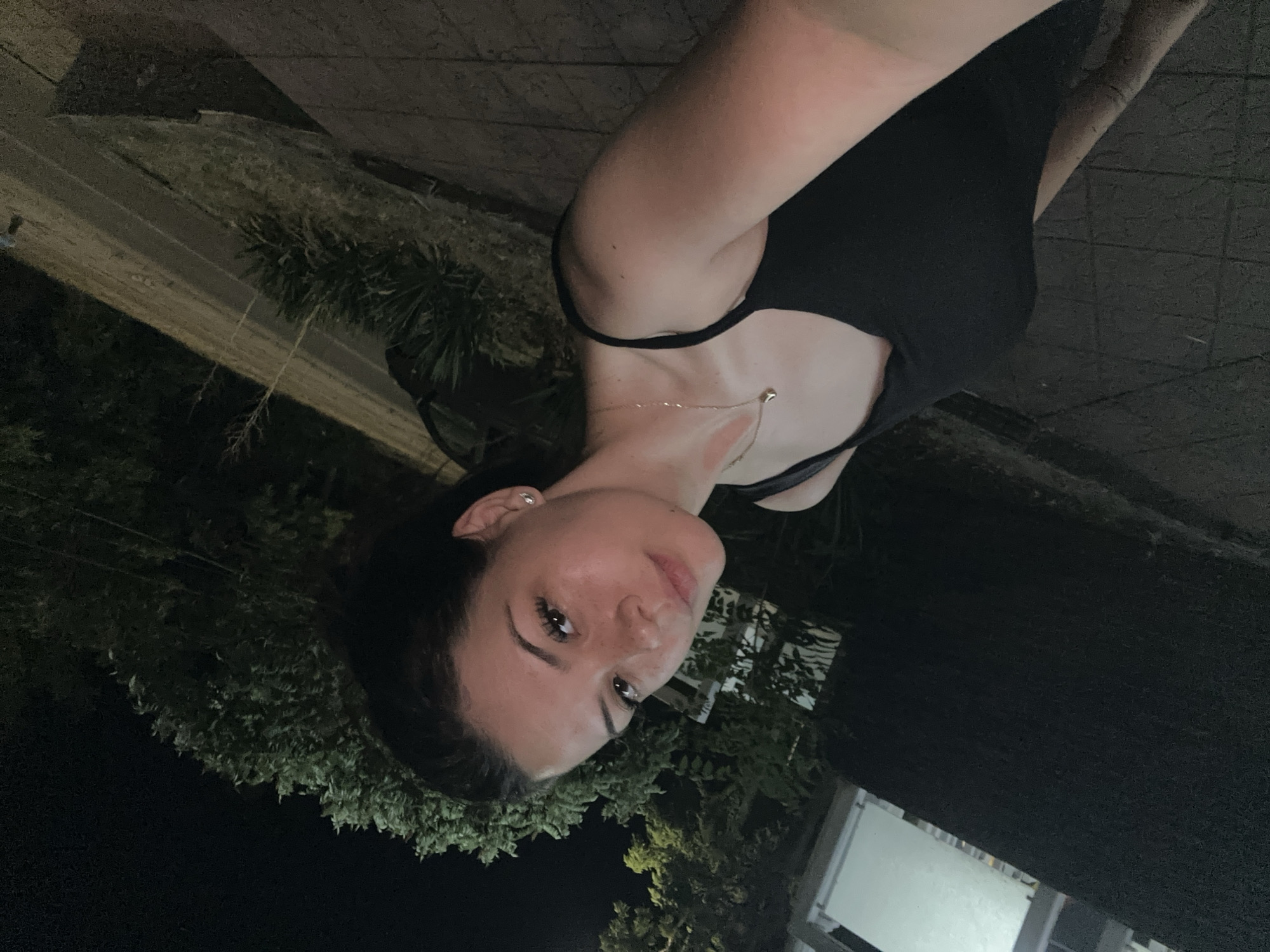 A young woman with dark hair and freckles poses for a selfie outdoors at night. She is wearing a black tank top and a delicate necklace. The background is dark, with some trees and a hint of a building. The setting appears to be an urban area.