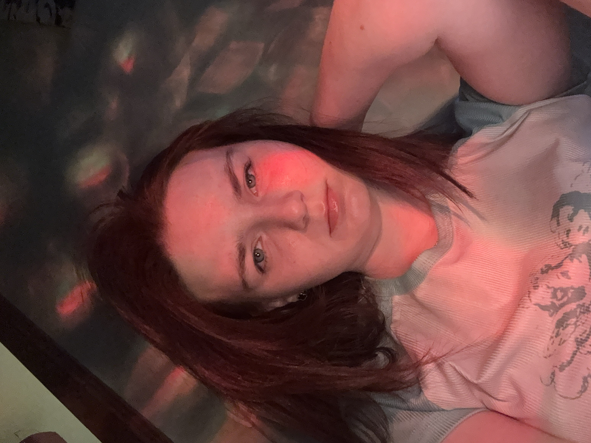 The media file is a portrait of a person, likely a young adult female, lying down and looking directly at the camera. Her head is angled to the left, and her dark brown hair is spread out. Her face is illuminated primarily by a strong reddish-pink light, which casts a glow on her left cheek and lips. Her eyes are light-colored, appearing blue or green, and she has a neutral, calm expression.
She is wearing a light-colored, possibly light blue or grey, ribbed t-shirt with a crew neck. A faint, indistinct graphic design is visible on the right side of her shirt (viewer's left), but no specific imagery or text can be clearly discerned.
The background is dark and out of focus, featuring several blurred, colorful light projections or reflections in shades of red, pink, and green. These suggest patterned or dynamic lighting, possibly from a disco ball or similar light source, creating a moody, atmospheric setting. The overall impression is an indoor scene, likely in low light conditions, suggesting evening or night. The activity appears to be posing for a photograph in this particular lighting. The location context is Unknown, Russia. No discernible text is visible in the image.