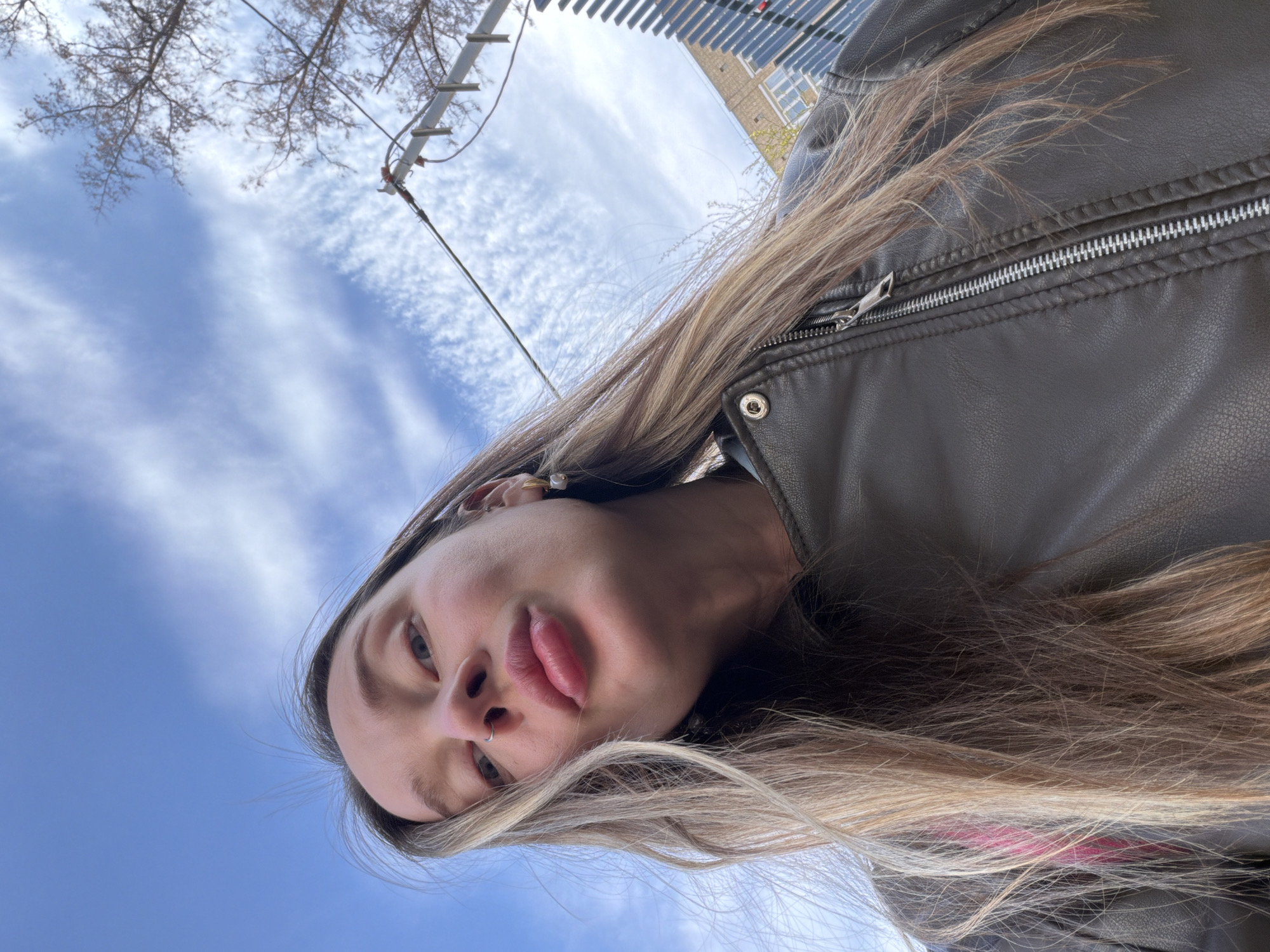 A low-angle close-up portrait of a young adult female. She has long blonde hair, facial piercings (including a nose ring), and is wearing a brown leather jacket. The background consists of a blue sky with scattered clouds. Above her, there is a utility pole with branches. The photo could be Moscow, Russia.