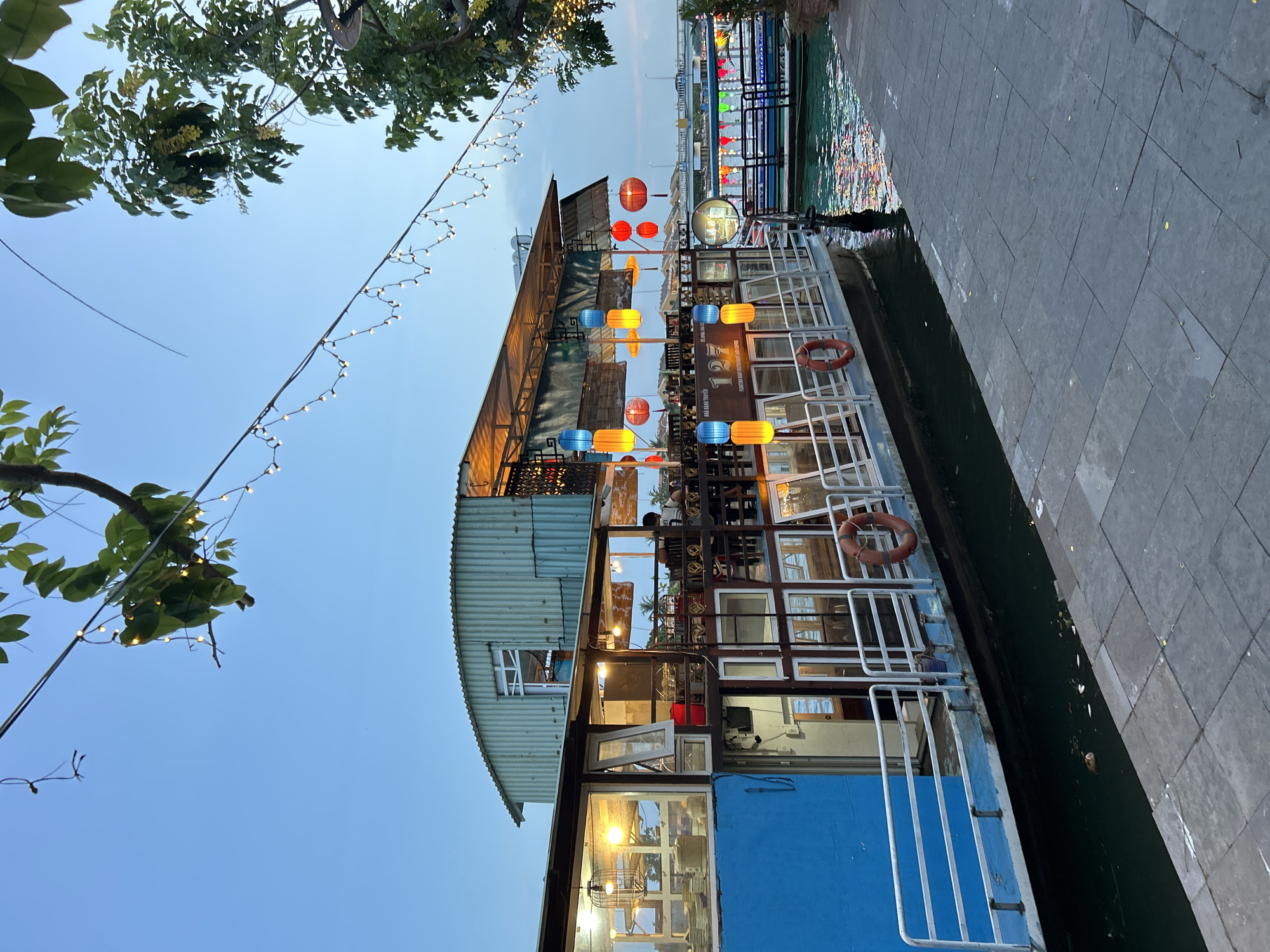 A floating restaurant and coffee shop, identified by the signage "127 FLOATING RESTAURANT AND COFFEE," is docked alongside a paved walkway. The vessel has a blue hull and a corrugated metal upper structure. Ornate lanterns, colored in blue, yellow, and red, hang from the roof of the restaurant. String lights are suspended over the walkway and through trees. The water reflects the colorful lights of the boat. In the background, a bridge spans the waterway, and other boats are visible. The sky is a soft blue, suggesting dusk or early evening. There is a single person visible sitting at a table inside the restaurant. A life preserver is attached to the railing of the boat.