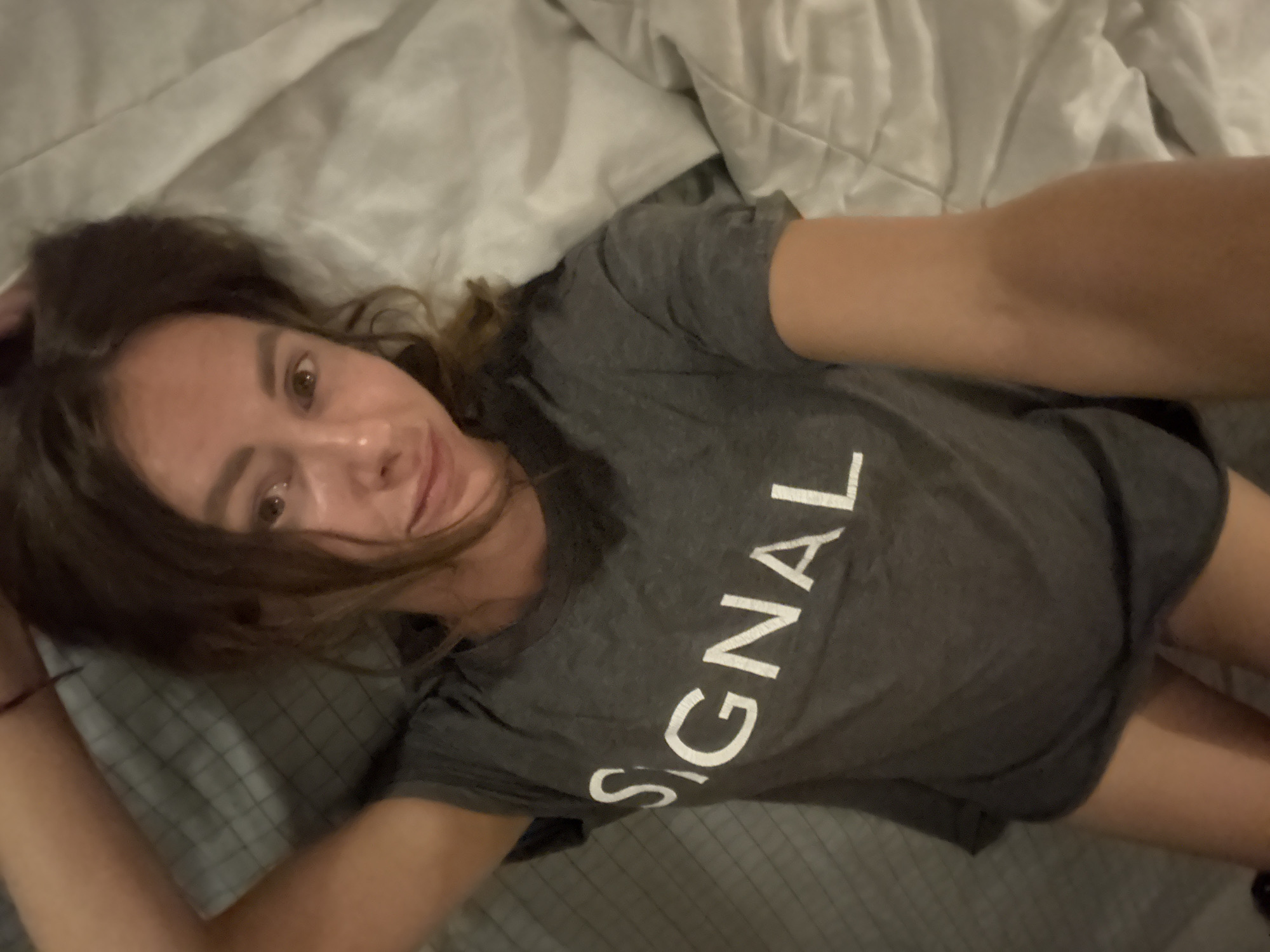 A woman lies on a bed, wearing a gray t-shirt with the word "SIGNAL" in white lettering. She is facing the camera, with her head resting on a gray pillow. The location is possibly in North Kuta, Indonesia.