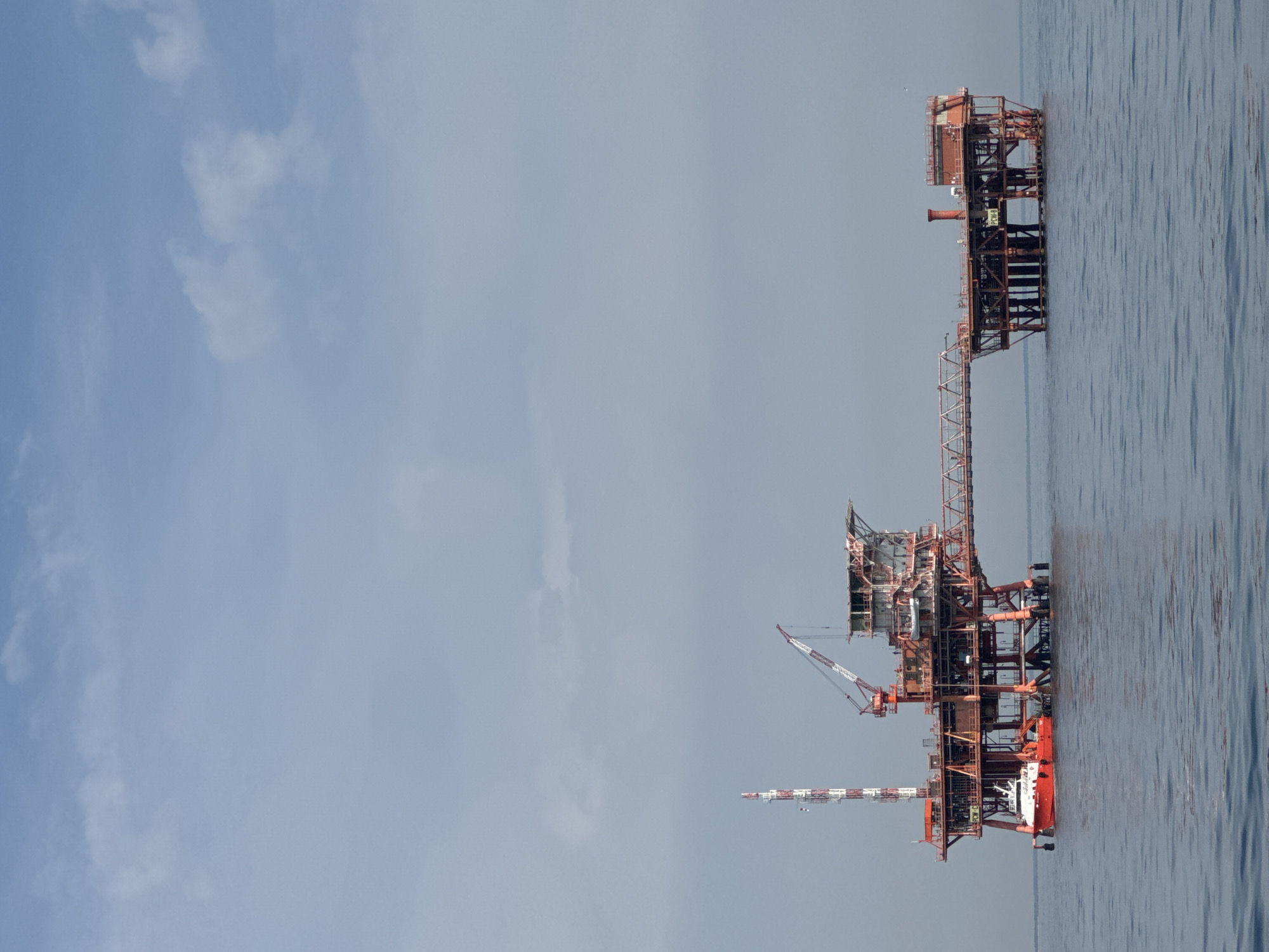 An offshore oil rig, painted predominantly orange and red, stands in the calm, blue water of the Adriatic Sea. The rig is connected by a long, metallic walkway to a smaller platform on the right. A red and white boat is docked to the left side of the main platform. The sky above is a pale blue with scattered, wispy clouds. The oil rig is located in the waters off the coast of Marina di Ravenna, Italy. There are no people visible in the image.