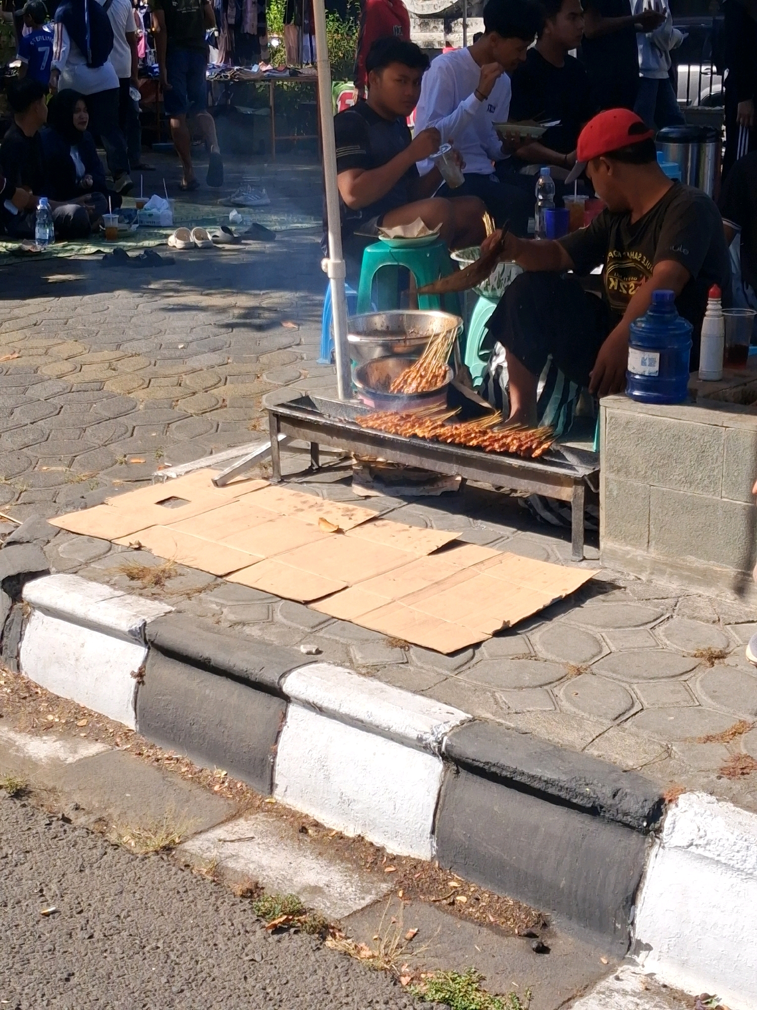 In the city of Surakarta, Indonesia, on a sunny day, a street food vendor prepares satay outdoors. The scene depicts a section of a paved walkway bordering a street. The walkway is constructed of interlocking grey concrete pavers. A curb separates the walkway from the asphalt street, with the curb painted in alternating black and white sections.
The vendor, a man wearing a red cap and a dark t-shirt, is seated on a green plastic stool. He is tending to a charcoal grill filled with satay skewers. More satay is arranged on a metal rack in front of the grill, glistening from the heat. Nearby, a large blue water dispenser, a stainless steel pot, and various cups and condiments are visible.
Two young men are seated on the ground to the left of the vendor. One, wearing a black t-shirt, is looking at the vendor. The other, in a white long-sleeved shirt, is holding a bowl and a drink. Several people are standing and walking in the background, suggesting a busy marketplace or street. One person, partially visible, wears a blue shirt with a white number "7" on it. The general atmosphere is casual and appears to be a typical street scene in Surakarta.