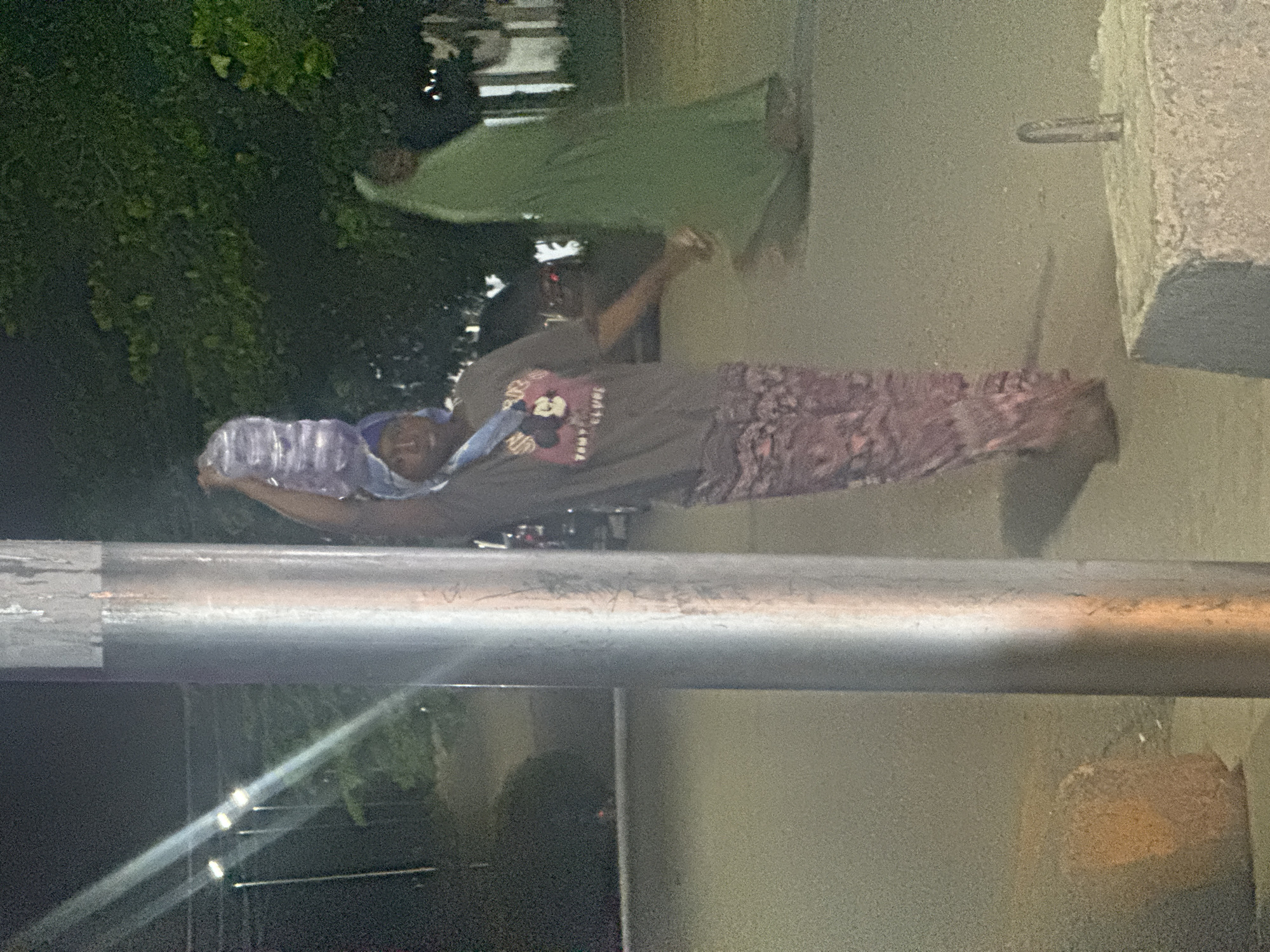 A person walks down a street in Abuja, Nigeria, balancing a stack of bottled water on their head. The person is wearing a grey t-shirt with a graphic print and a patterned skirt. To the right, another person in a green hijab walks away from the camera. The scene appears to be at dusk or night, with artificial lighting visible in the background. A metal pole is prominent in the foreground, and a concrete structure with a metal fixture is in the lower right corner.