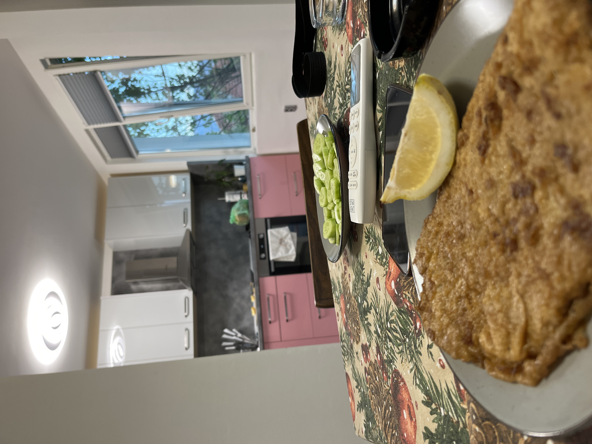 The scene depicts a table setting in an interior space, possibly a dining area, likely located in Karavelovo, Bulgaria. The table is covered with a patterned tablecloth, featuring pine cones and greenery. On the table is a plate with a fried food item and a lemon wedge. A small dish contains sliced cucumbers. Adjacent to the plate are a smartphone, remote control, and headphones. In the background, there is a view of a modern kitchen with white and pink cabinets. A window is visible above the cabinets, showing greenery outside. A circular light fixture is mounted on the wall.