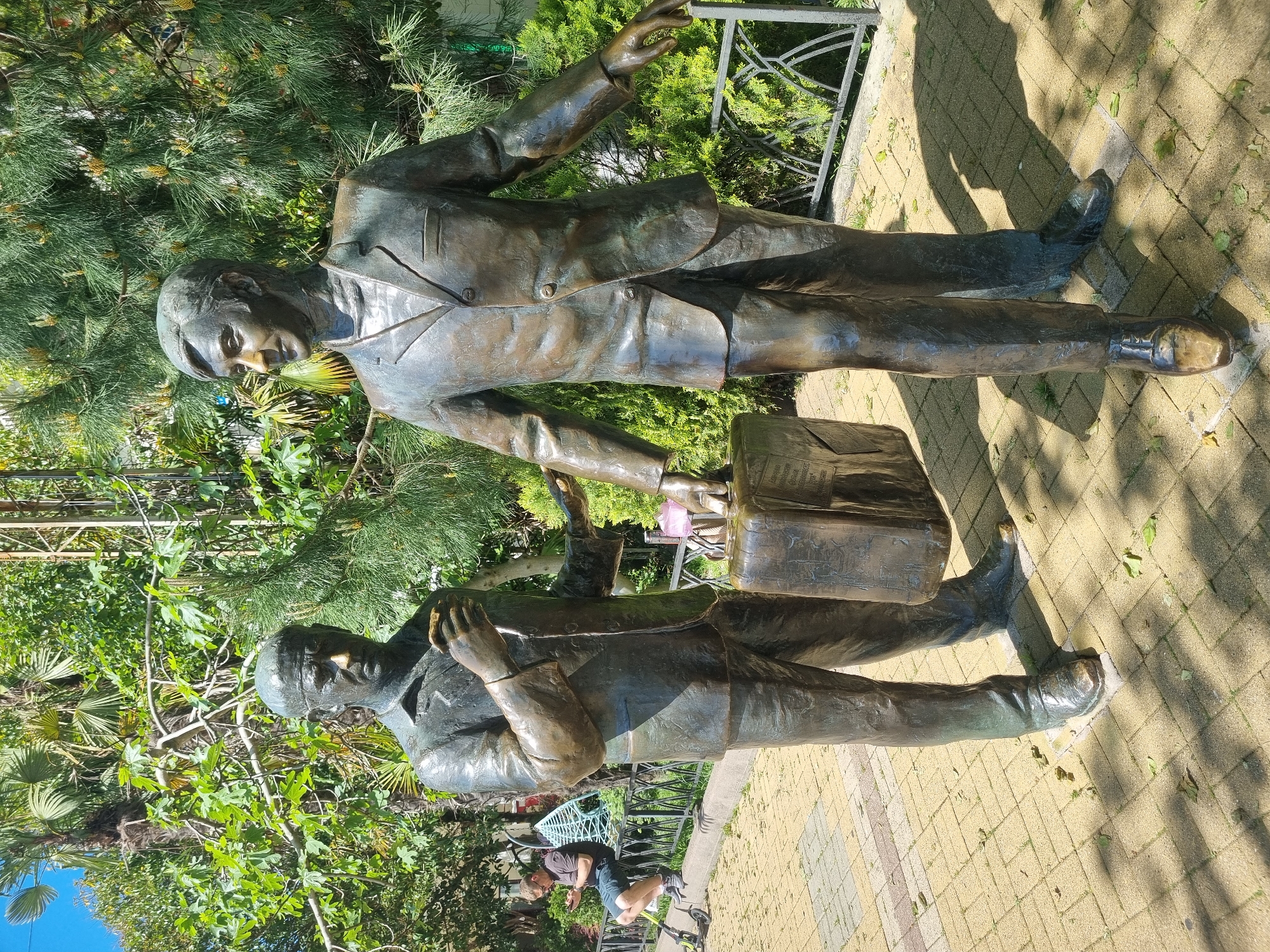 The image shows a bronze sculpture of two men in suits standing close to each other in an outdoor setting. One man is holding a briefcase, while the other has his arm extended in a gesture. The background consists of trees and foliage, with a paved walkway beneath the statues. The location is the Kurortny Avenue in Sochi, Russia. A person is seen sitting in the background.