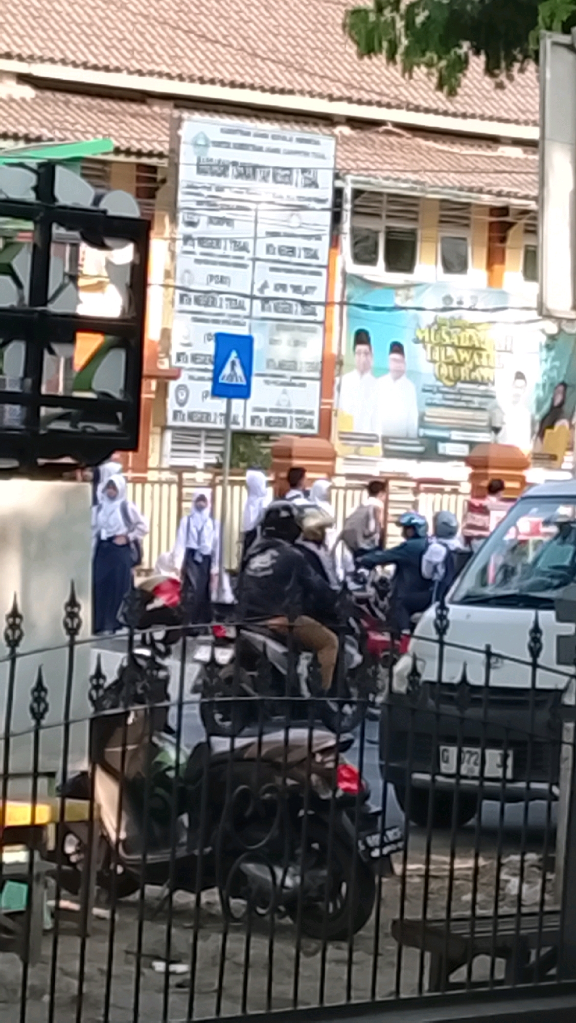 The image depicts a street scene in Slawi, Indonesia, with a focus on a group of individuals, many of whom appear to be students in uniform. In the foreground, a black metal fence obscures the lower portion of the scene, behind which several motorcycles are parked. One motorcycle is prominent, with a rider wearing a black jacket and a light-colored helmet. The background features a building with a tiled roof and windows, and a large white sign with text in Indonesian, likely an informational board or advertisement, is mounted on the building's facade. To the right of this sign, another banner or poster with images of men in traditional attire and Indonesian text is visible. Several individuals wearing white shirts and dark skirts or pants, characteristic of Indonesian school uniforms, are standing and walking in the middle ground, some carrying backpacks. The overall impression is one of a busy street near a school or educational institution, possibly during a time of transition such as morning arrival or afternoon dismissal. The lighting suggests it is daytime, with sunlight casting shadows.