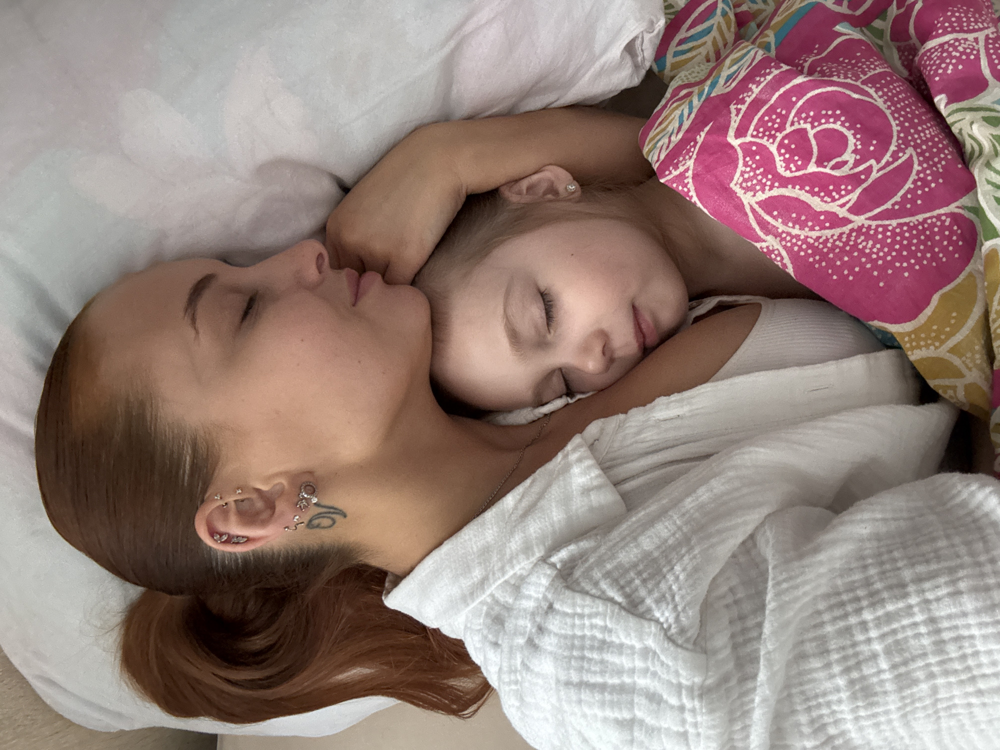 A young woman and a child are lying down together in a bed with white sheets. The woman has auburn hair pulled back in a low ponytail, multiple piercings in her left ear, and a tattoo near her ear. She is wearing a white textured shirt and a silver chain necklace. The child, partially covered by a pink floral blanket, is wearing a white tank top. Both are lying on their sides with their eyes closed. The location is inside a building in Chastyye, Russia.
