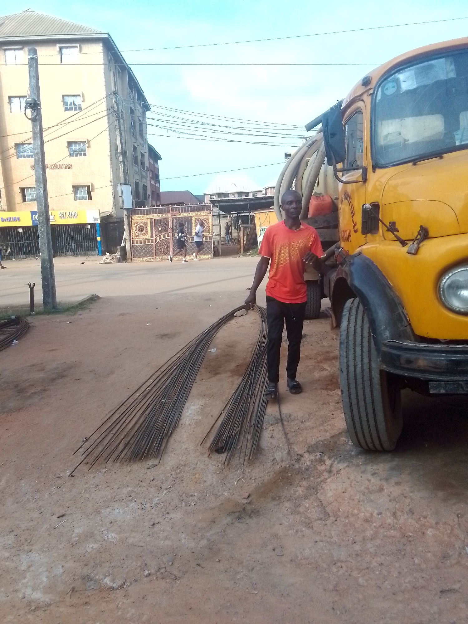 The image depicts a man standing on a dirt road in Nkpor, Nigeria. He is wearing an orange t-shirt and black pants, and he is holding two bundles of long, thin metal rods. Next to him is a large yellow truck with hoses attached to its side. In the background, there is a multi-story building and a metal gate. Two people are visible in the distance near the gate. A building with the words "Betking" on it is also visible in the background. The road appears to be unpaved and dusty.