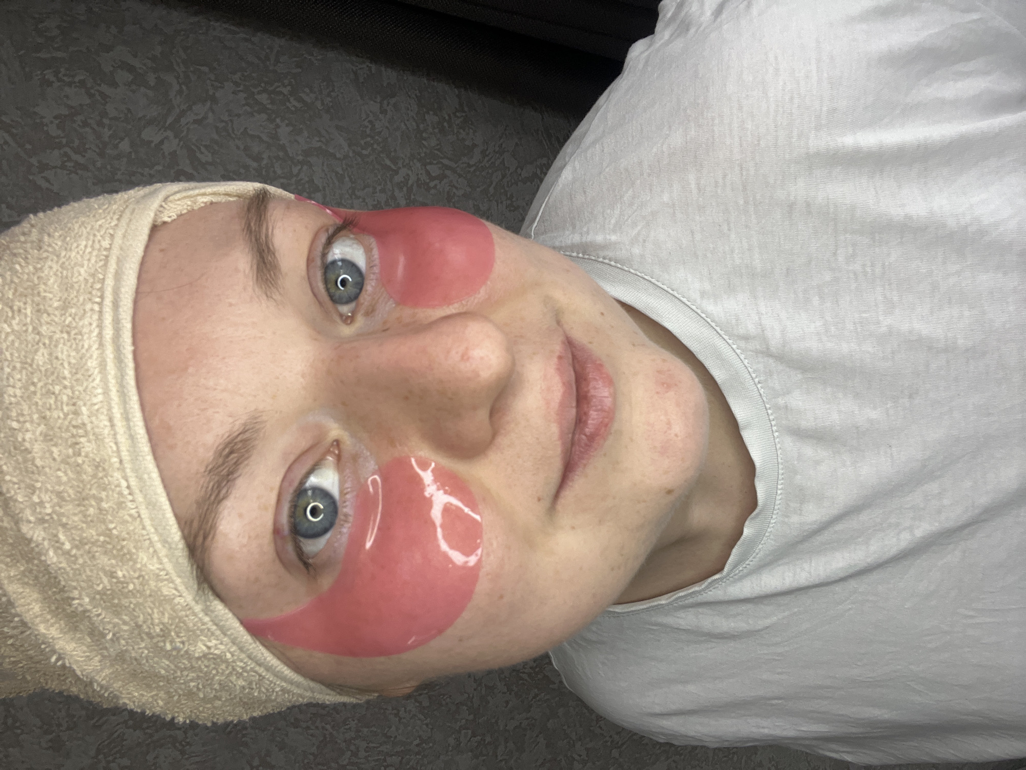 The image shows a woman with fair skin and blue eyes, wearing pink eye patches below her eyes and a towel headband. She is wearing a white t-shirt and appears to be indoors against a textured, gray background. The photo seems to be taken from a slightly low angle and rotated to the side. There's no indication that the image was taken in Раздолье, Russia.