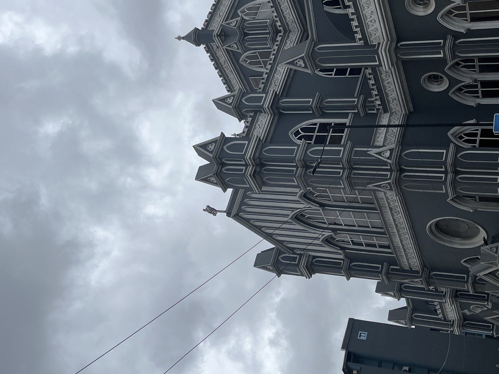 The image shows a gray building with intricate white trim under a cloudy sky. The building appears to be a church or cathedral with Gothic architectural details, including arched windows and spires. Power lines are visible in the foreground. A smaller, modern building is also visible adjacent to the older structure. This is St. Timothy's Anglican Church in Lagos, Nigeria.