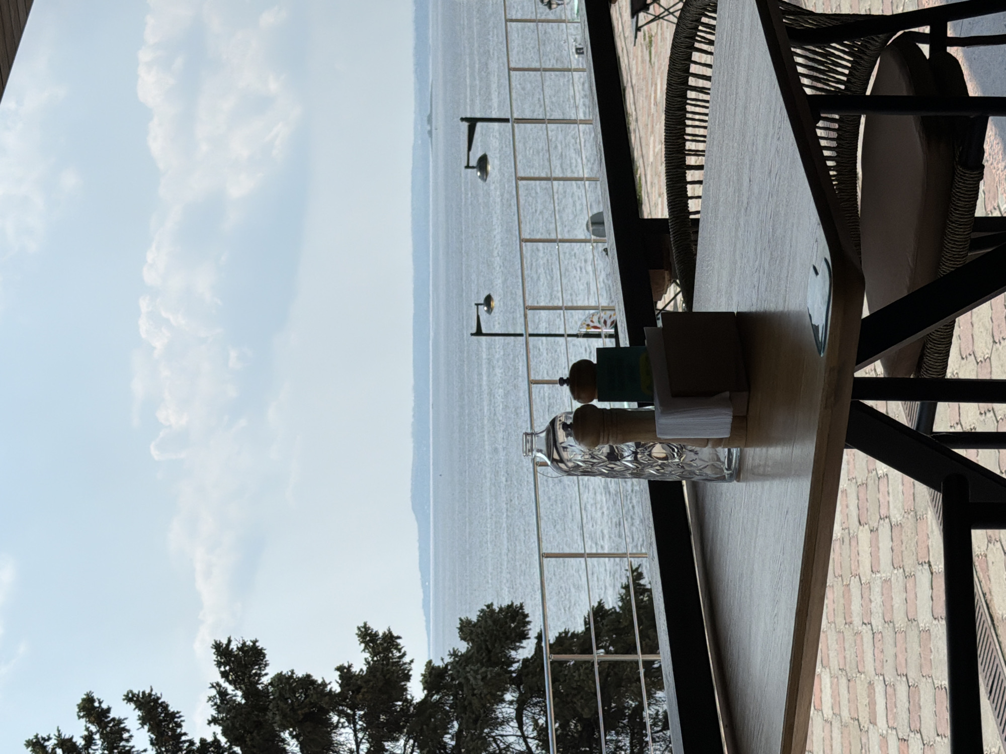The image shows an outdoor seating area overlooking a large body of water, likely the Volga River, in Saratov, Russia. In the foreground, a wooden table is set with a glass bottle, salt and pepper shakers, and a napkin holder. A wicker chair with a cushion is placed at the table. The background features a metal railing and two black lampposts, with a tree on the left side of the frame. The water is calm, and a distant shoreline is visible under a cloudy sky. The paving stones beneath the table and chairs suggest a well-maintained outdoor space, possibly a restaurant or cafe.