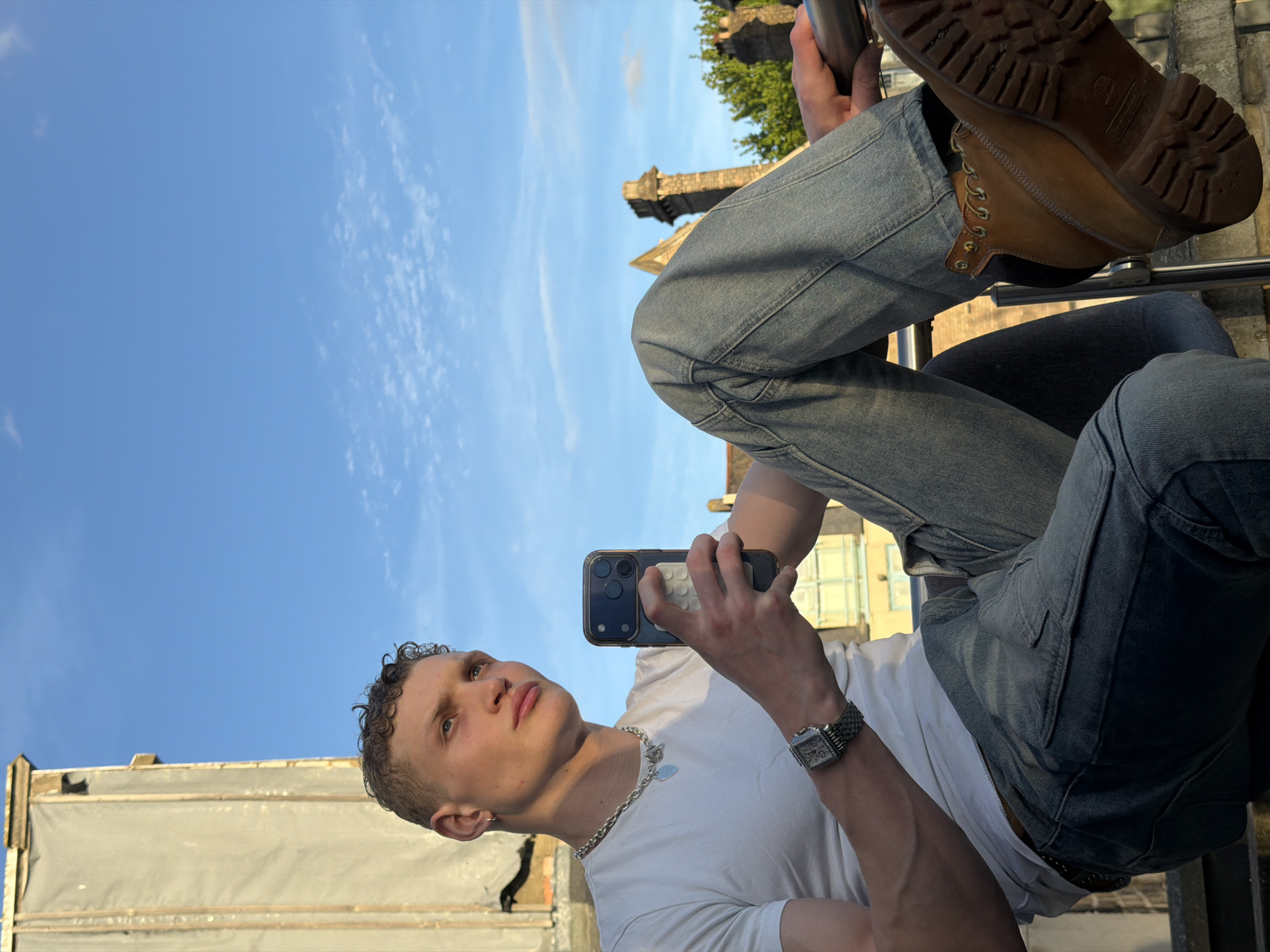 A medium-shot, rotated image showing a young man sitting and taking a selfie in an urban location. He is wearing a white t-shirt, light blue denim jeans, and brown work boots. The man has fair skin, short curly hair, and is wearing a silver chain necklace and a rectangular-faced wristwatch. He is holding a smartphone in his hands, looking up and away from the camera.
Behind the man and angled towards the sky is architecture: the top of an old stone tower, part of a white building with a weathered facade, and hints of greenery. The background reveals a bright blue sky with light clouds. The scene appears to be in an elevated outdoor location with railings. The overall impression is that the man is in a city location, but I cannot confirm it is in the city of London in the United Kingdom.
