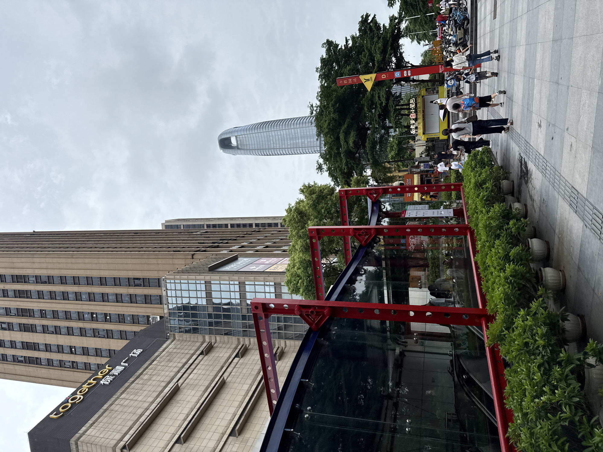 The image depicts a street scene in Haizhu District, China. In the foreground, a row of potted green plants lines a paved walkway. A modern building complex with a glass and red metal structure is visible on the left, with an escalator descending into the ground level. Behind this, a tall, brown skyscraper with numerous windows dominates the skyline, bearing a "together" logo and Chinese characters. To the right, a distinctive, curved, glass skyscraper stands in the distance. Several people are walking on the sidewalk. A yellow food cart is parked on the right side of the street, and several motorcycles are parked nearby. The sky is overcast.