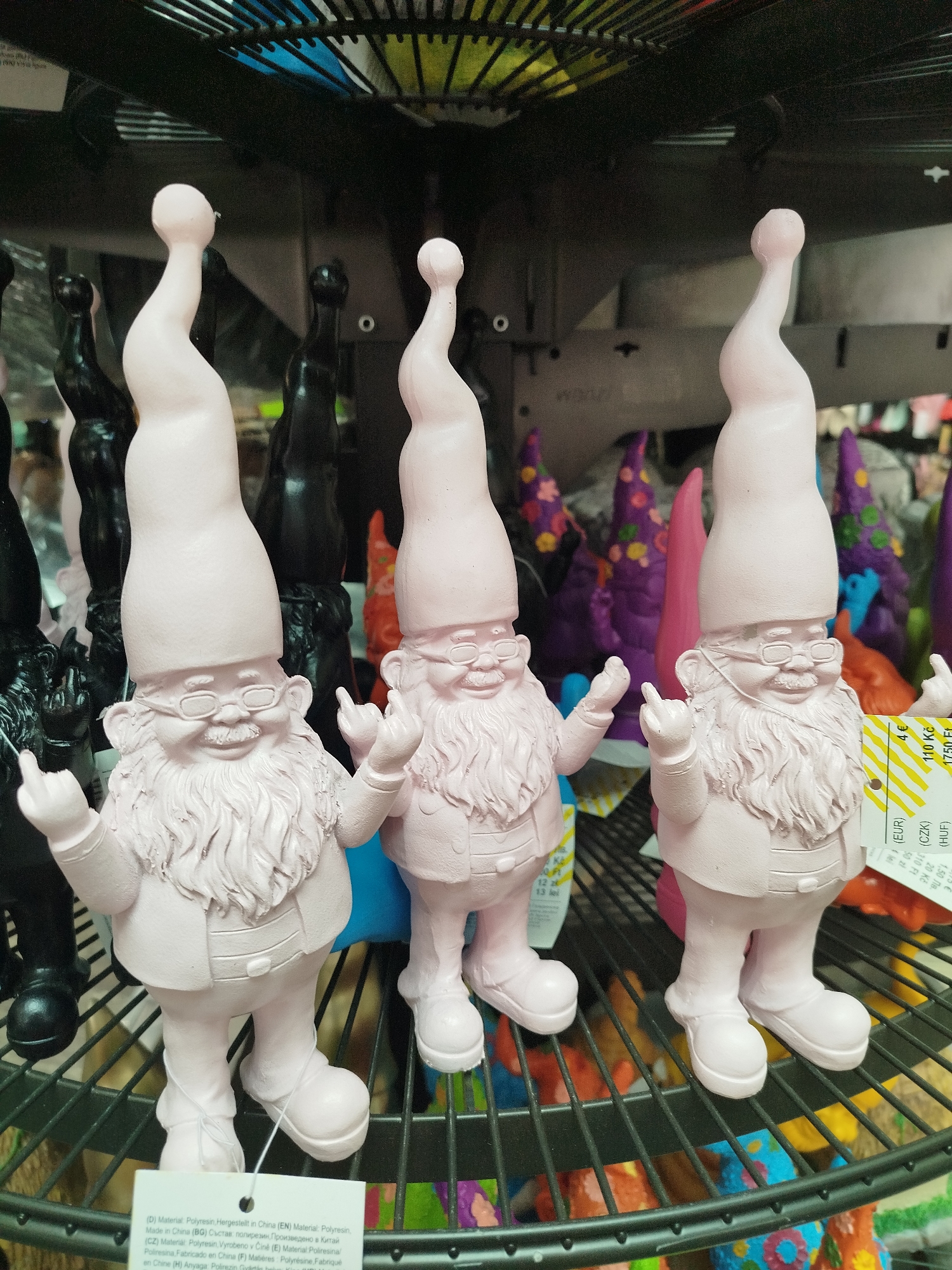 Three pink garden gnomes with long pointed hats, beards, and glasses stand on a black metal shelf. The gnome on the left has its middle and index fingers raised, while the other two gnomes have their middle fingers raised. The gnomes are positioned in the foreground, with other gnomes and colorful figures visible on shelves behind them. The background consists of shelves with various decorative items. The image appears to have been taken in a retail store. The location is identified as Offenbach am Main, Germany.