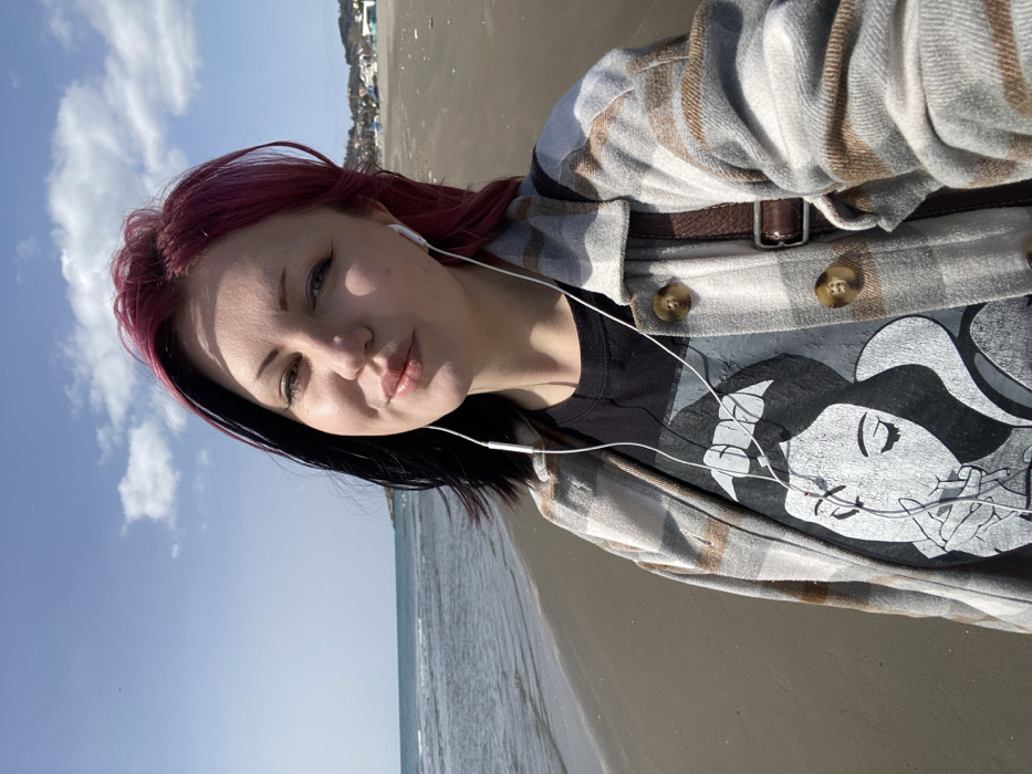 An individual is depicted in a close-up, selfie-style photograph taken on a sandy beach in Ulcinj, Montenegro. The subject is a female, appearing to be in her mid-20s to early 30s, with a two-toned hairstyle featuring black hair on the left side and magenta-red hair on the right. She is looking directly at the camera with a slight smile and pursed lips, wearing white wired in-ear headphones. Her attire consists of a black t-shirt featuring a white graphic print of a woman, possibly Snow White, with closed eyes and a hand near her mouth, layered under an open light gray and brown plaid shirt or jacket with brown buttons. A brown leather bag strap is visible over her right shoulder.
The background reveals a wide, calm body of water, likely the Adriatic Sea, on the left, with gentle waves reaching the shoreline. The sandy beach extends into the distance on the right, where distant low-rise buildings and a treeline are visible along the coastline, indicative of a coastal settlement. The sky is clear blue with scattered white cumulus clouds. The scene is illuminated by natural sunlight, suggesting a bright day.
