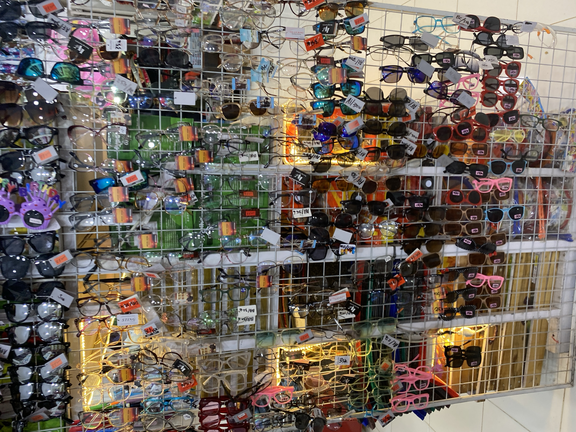 A retail display unit, likely situated in a market or shop in Lamteumen Barat, Indonesia, showcases a diverse assortment of eyewear. The unit is constructed from a white wire grid system, organized into multiple sections, each densely packed with sunglasses and eyeglasses. The eyewear varies in style, color, and frame material, with many items featuring colorful lenses and decorative elements. Small price tags or labels are visible on many of the glasses, indicating they are for sale. The overall impression is of a bustling marketplace offering a wide selection of affordable eyewear.