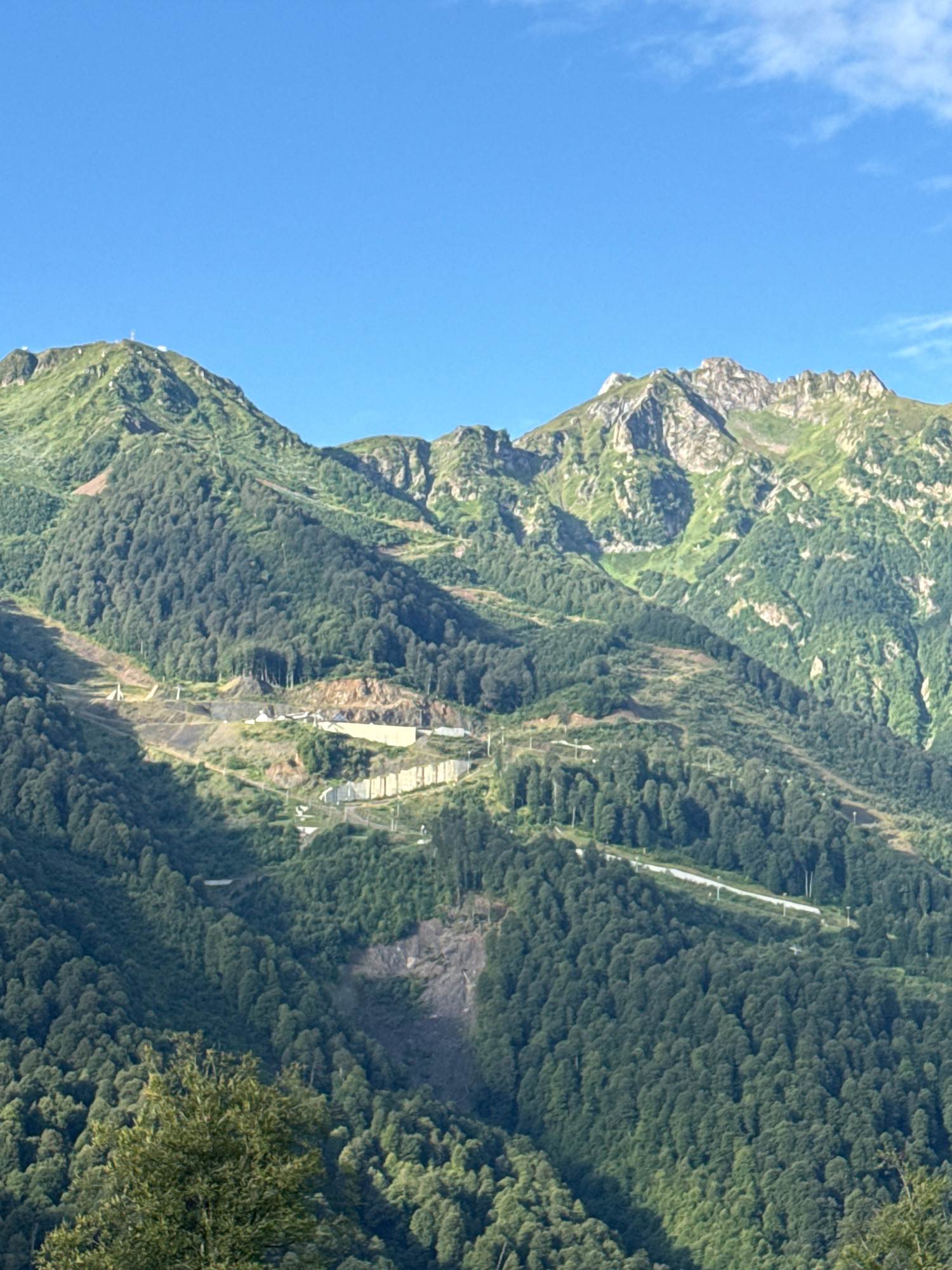 The image depicts a mountainous landscape with lush green forests and rocky peaks under a clear blue sky. In the foreground, a dense forest covers the lower slopes. As the view moves up the mountains, clearings and man-made structures become visible. These include what appear to be ski slopes or pathways carved into the mountainside, with supporting structures like ski lifts or pylons. Some areas show evidence of construction or excavation, with exposed rock and soil. The overall scene suggests a resort or recreational area situated in a natural mountain environment. This location is identified as being in Estosadok, Russia, a resort town known for its proximity to the Caucasus Mountains and its winter sports facilities.