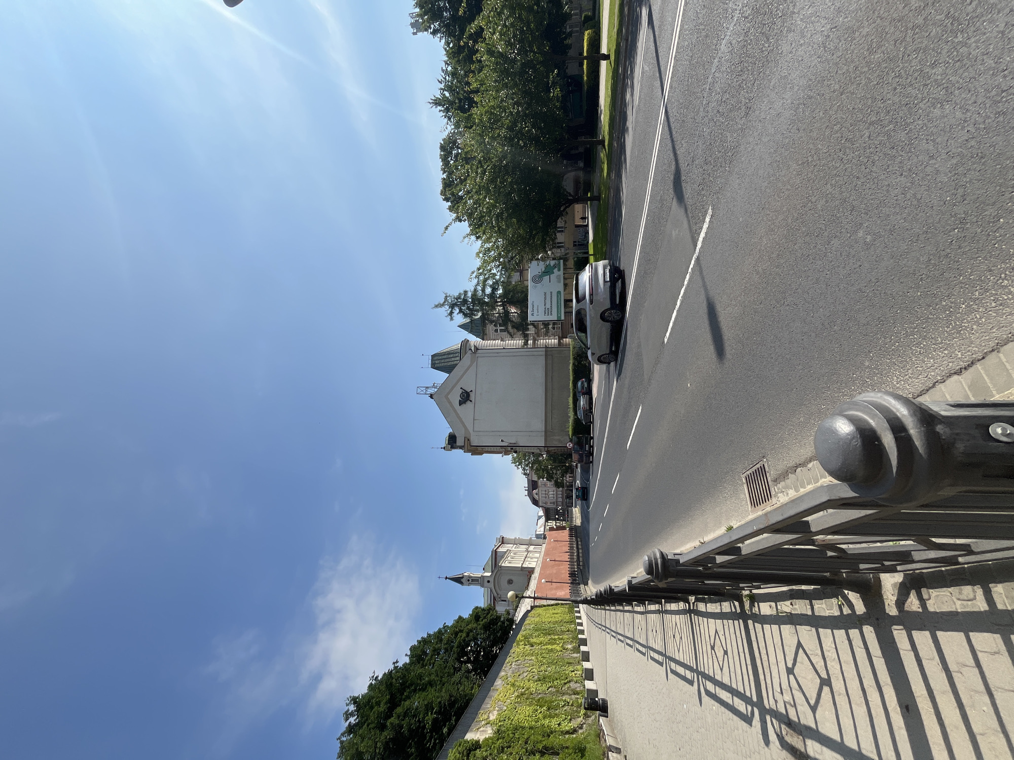 The image shows a street scene in a city. On the left side of the image is a sidewalk with a black metal fence. Behind the fence is a wall covered in green ivy. Further behind the wall, a building with a tall tower and a cross on top is visible. The street in the center of the image is paved and has white lane markings. Several cars are driving on the street, including a silver SUV. On the right side of the street, there are trees and buildings. The sky is blue with a few white clouds. The scene appears to be a sunny day.