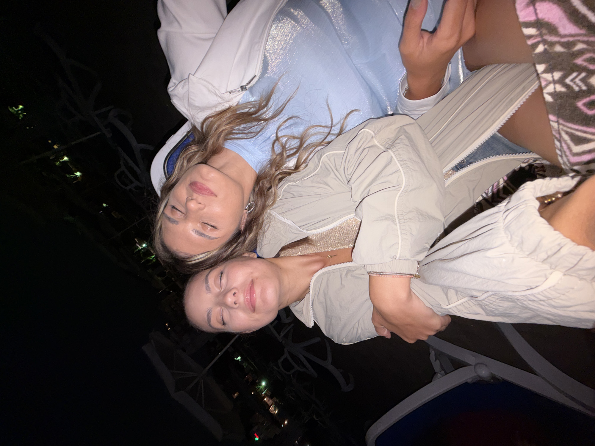 The image depicts two young women with their eyes closed, appearing relaxed or asleep, at night on a beach in Lozenets, Bulgaria. The woman on the left is leaning against the woman on the right, who has her head resting on the left woman's shoulder. Both are wearing casual clothing. The background shows dimly lit beach chairs and umbrellas, suggesting a resort area. The lighting is low, indicating it's after sunset, and the overall mood is peaceful.