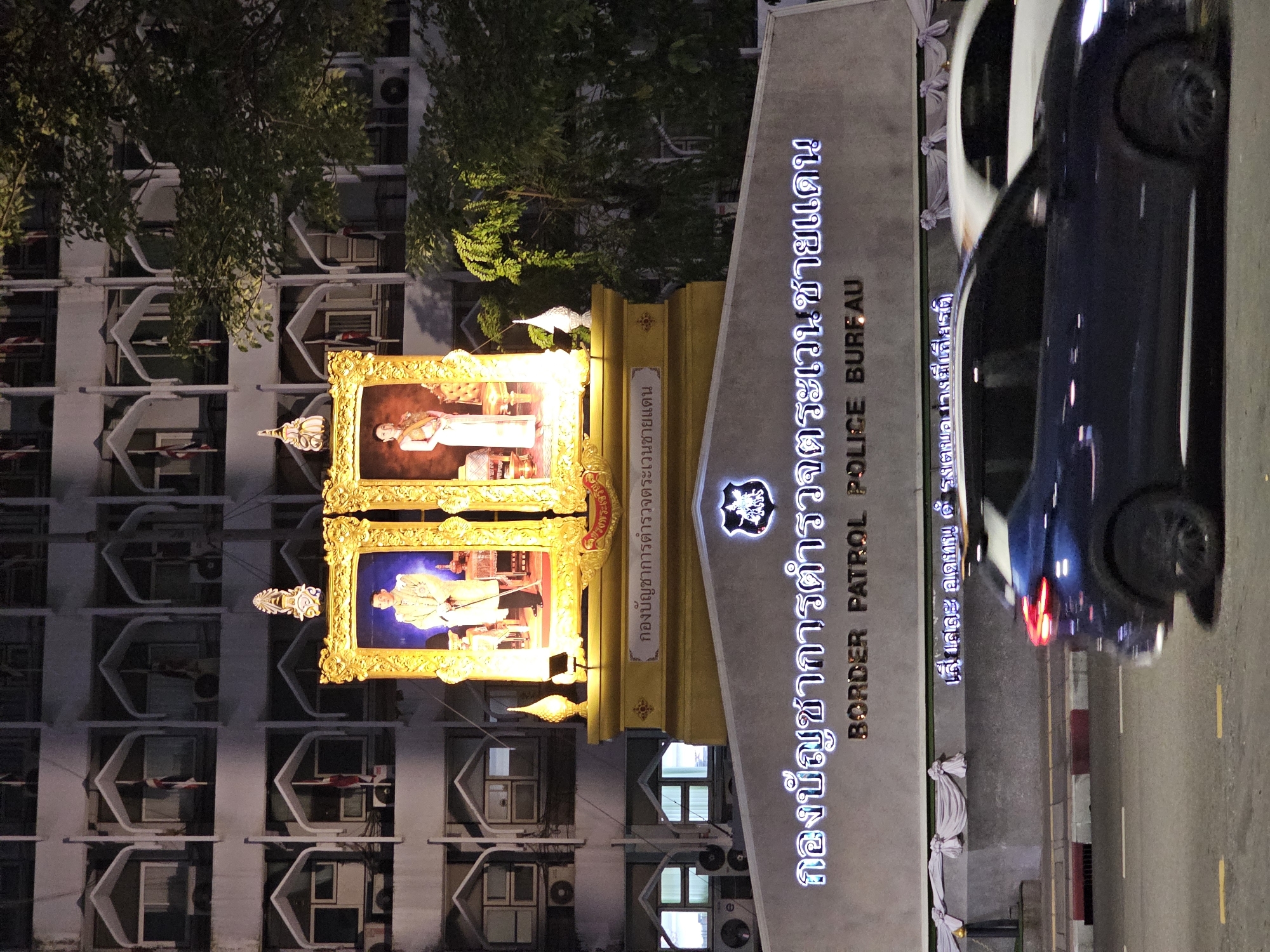 The image depicts the exterior of a government building at night, likely in Bangkok, Thailand, identified by the sign reading "BORDER PATROL POLICE BURFAU". In the foreground, a dark blue SUV drives past on a paved road. Behind the road, a grey wall is adorned with white draped fabric and a crest above a prominent sign with Thai lettering and the English translation. Further back, a two-tiered golden structure displays two framed portraits, one of a man in traditional attire and another of a woman in similar dress. Behind this display, the facade of a multi-story building is visible, characterized by numerous arched windows and balconies, some of which have small flags. Lush green trees are present to the right of the building. The overall scene is illuminated by artificial lighting.