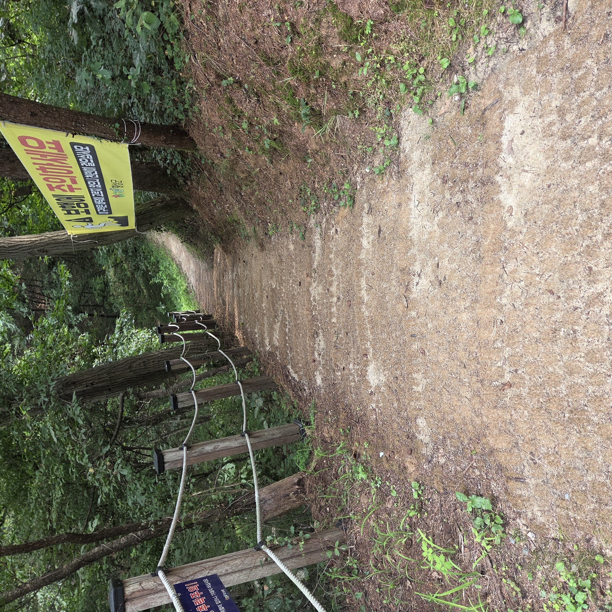 A dirt path with shallow steps descends through a wooded area. A rope fence with wooden posts lines the left side of the path. A yellow banner with Korean text is attached to a tree on the right side of the path, and a blue sign with Korean text is attached to a post on the left. The path is surrounded by trees and foliage. The location is identified as Gapcheon-myeon, South Korea, based on the text on the banner.
