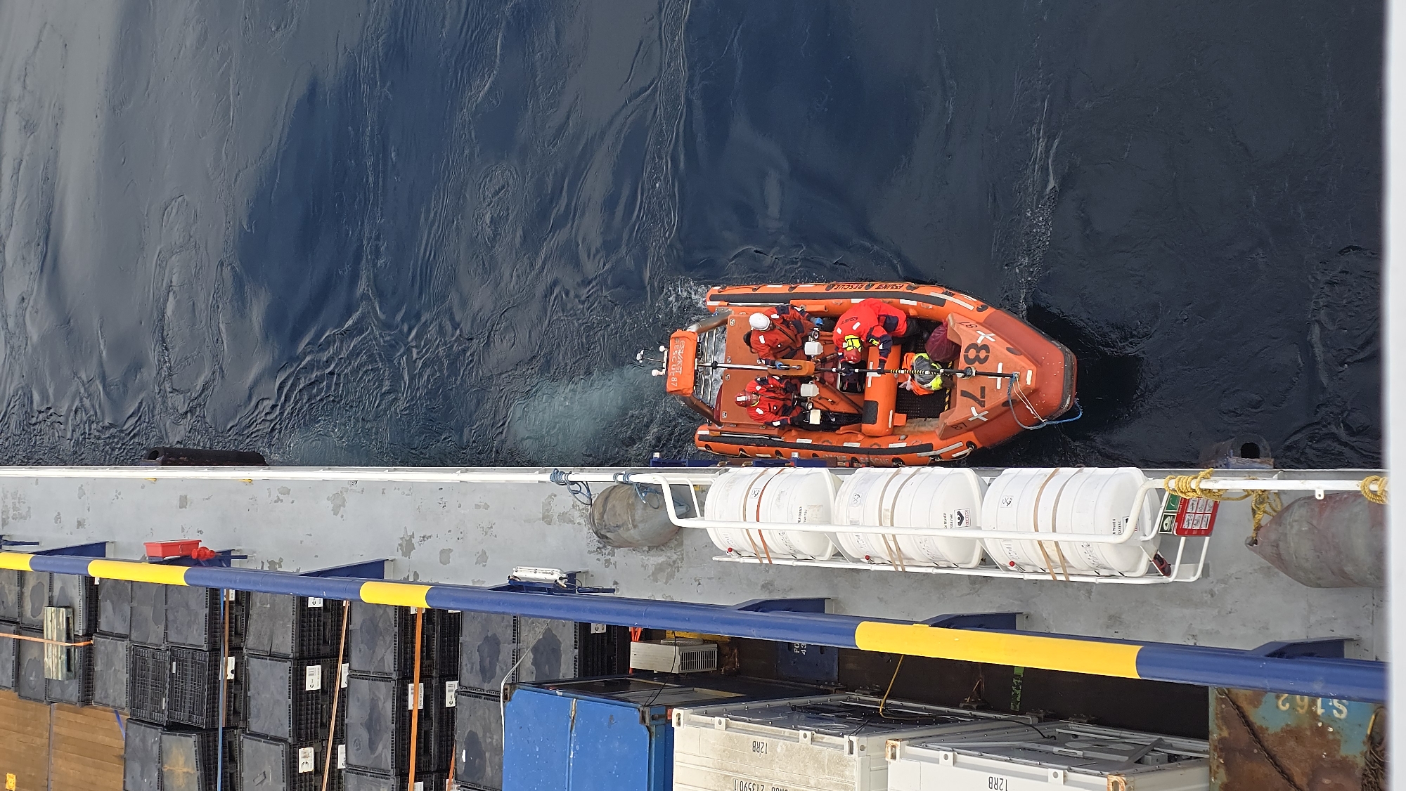 A high-angle shot shows an orange rigid inflatable boat (RIB) underway next to a vessel. The RIB contains at least four individuals wearing red survival suits. The vessel adjacent to the RIB has a gray deck with various items stored on it, including black plastic crates, white cylindrical containers held in a rack, and other containers. The sea is dark blue and slightly choppy. The location is at sea; specific city information is unavailable.