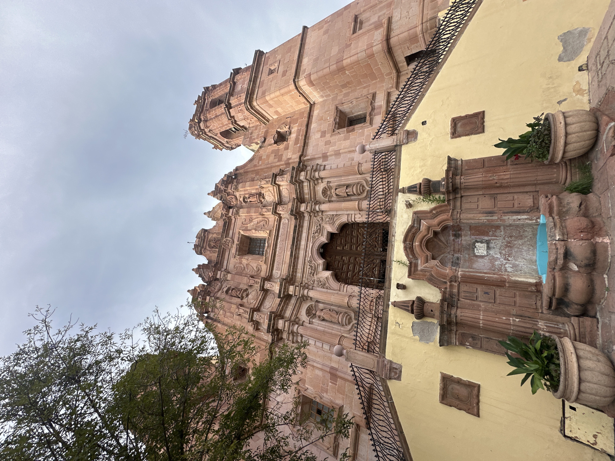 The image shows a low-angle view of the exterior of the Templo de San Francisco in Zacatecas City, Mexico. The church is constructed of pinkish-red stone and features an ornate Baroque facade with intricate carvings, statues, and columns. A large wooden door is centrally located within the facade. To the left of the main entrance, a green tree with dense foliage partially obscures the upper portion of the building. To the right, a bell tower rises above the main structure.
In the foreground, a wide, pale yellow staircase leads up to the church entrance. Black metal railings with decorative scrollwork line the sides of the stairs. At the base of the staircase, a fountain made of carved red stone is visible. The fountain has a central niche with what appears to be a blue basin. Two large, decorative planters with green plants are placed on either side of the fountain. The sky above is overcast with a pale, cloudy gray hue. The overall scene is bathed in soft, diffused light.