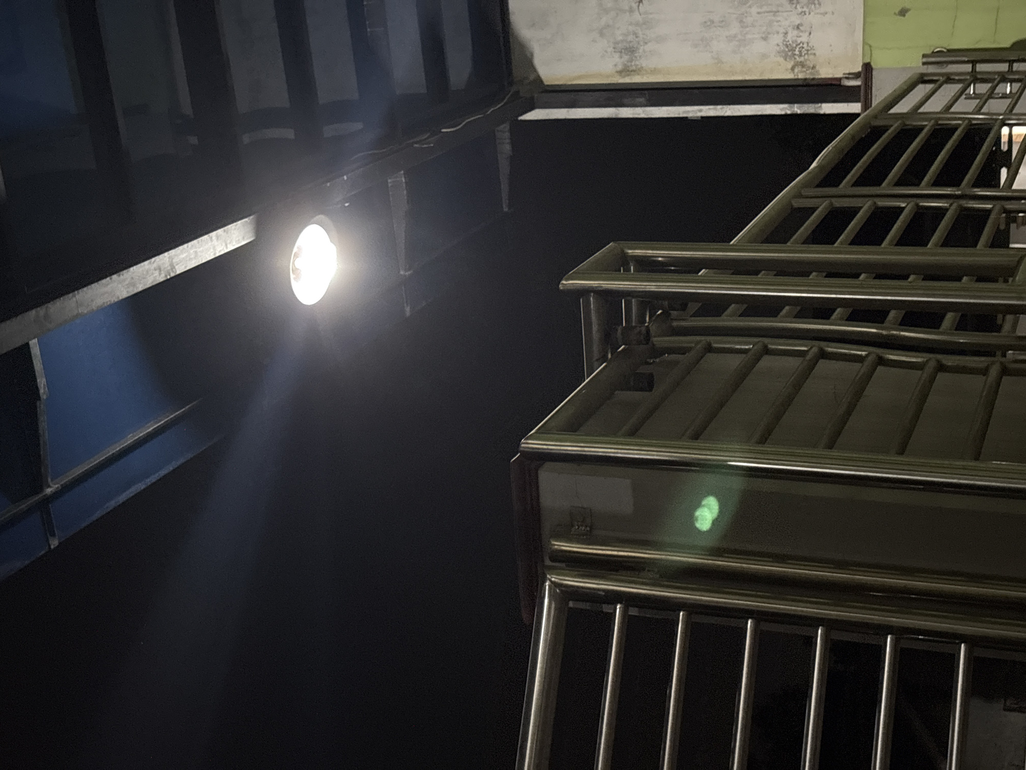 The image captures a nighttime scene on an elevated walkway or bridge in Gempol, Indonesia. The primary subject is a bright overhead light illuminating the area. In the foreground, a metallic railing system, likely part of the walkway's structure, is visible. This railing consists of horizontal bars supported by vertical posts. The railing is made of polished metal and reflects the ambient light.
To the right of the railing, a wall with a weathered, textured surface is present. It appears to be made of concrete or a similar material and has some discoloration and marks. Adjacent to this wall, a section of a green-painted surface is visible, suggesting the presence of another structure or element in the vicinity.
Above the walkway, a dark sky dominates the upper portion of the image. Several structural elements of the elevated walkway, possibly beams or roofing, are visible against the dark background. These structures appear to be made of dark-colored metal or concrete.
The overall impression is that of an outdoor public space at night, illuminated by artificial light. There are no people visible in the frame, and no specific actions or interactions are depicted. The environment appears to be urban or semi-urban, given the presence of the elevated walkway and constructed elements. The green painted surface hints at a possibly more brightly lit or colorful area nearby.