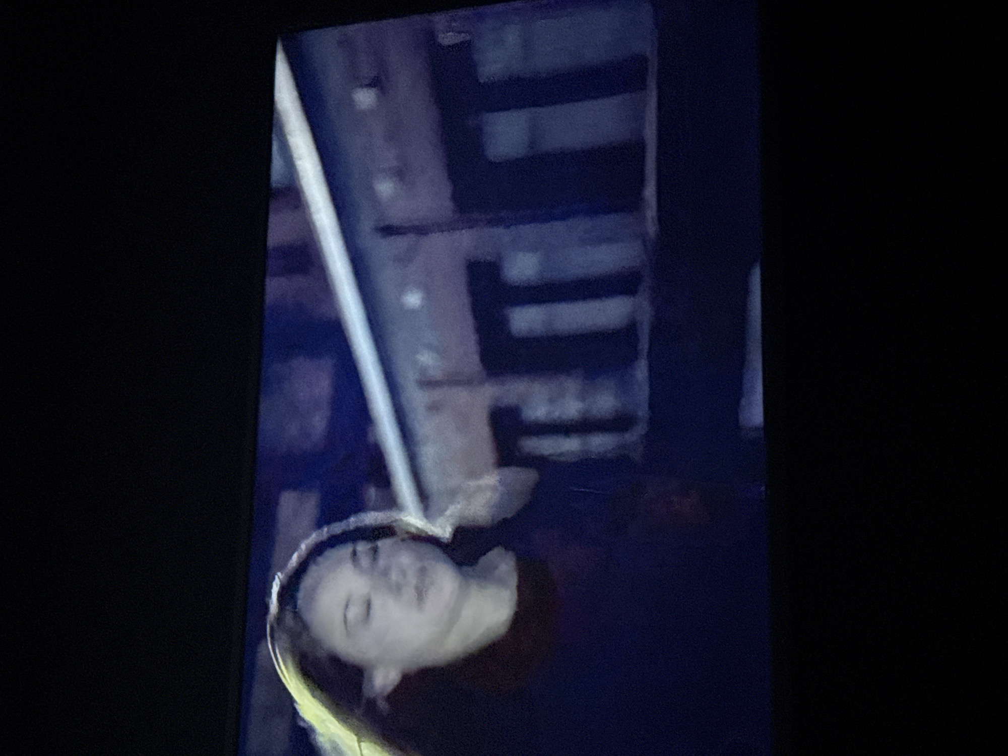 A still image from a screen displays an individual with light skin and hair, lying in what appears to be an urban environment. The face is visible and oriented horizontally to the left. Above the individual is a blurred architectural structure, possibly a building, with horizontal dark lines suggesting windows or ledges. The overall lighting is dim, with darker areas obscuring details. The background is largely dark, possibly indicating nighttime. It is impossible to determine the location is Mytishchi, Russia with the information presented in the image.