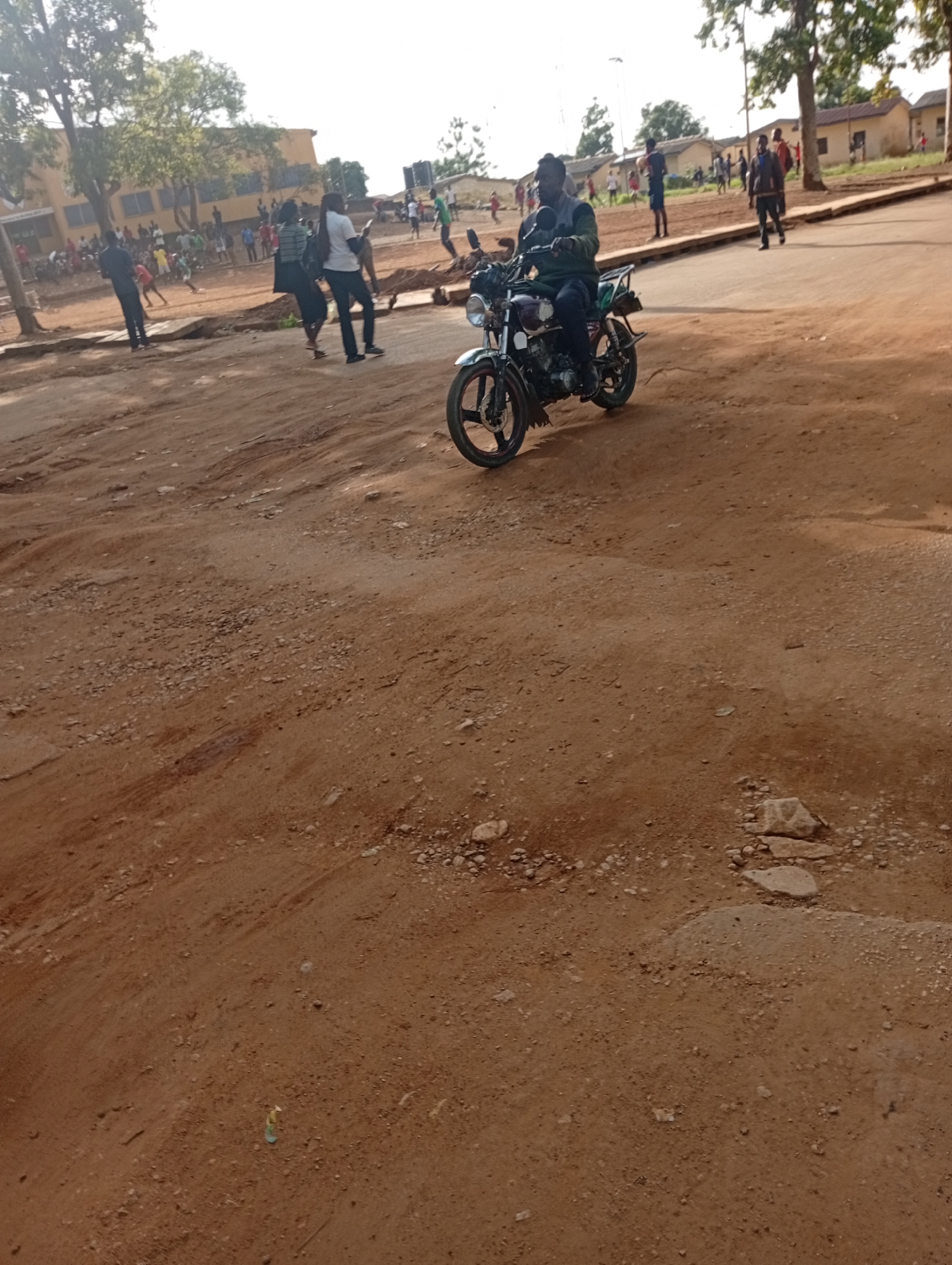 The image depicts an outdoor scene in Rafin Par, Nigeria. In the foreground, a dirt road with scattered rocks and tire tracks is visible. A man is riding a motorcycle on this road, wearing a dark jacket and a helmet. In the background, several people are gathered, and some appear to be playing or engaging in activities. Buildings with yellow walls and brown roofs are present, along with trees. The sky is overcast. The overall impression is of a busy community area with both people and vehicles present.