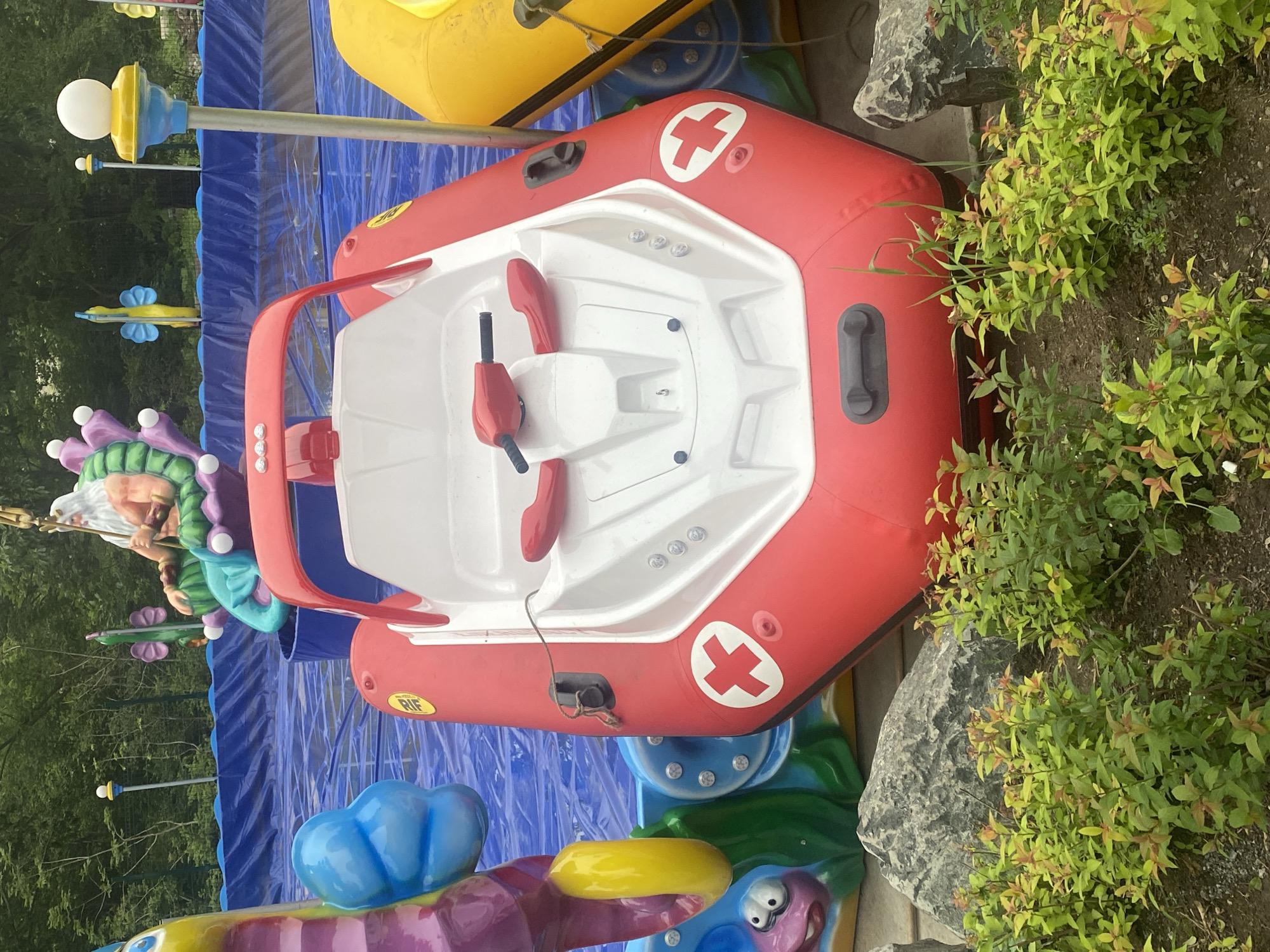 In an outdoor setting, a red and white inflatable boat designed to resemble a rescue vehicle sits in shallow blue water. The boat has a steering wheel and two seats. On its side is a white cross within a red circle, indicating a medical symbol. Behind the boat, a larger inflatable structure features colorful sea creatures, including a merman figure. The water is contained within a blue, tarp-like pool. Rocks and green shrubs are visible in the foreground. The background consists of trees and a fence.