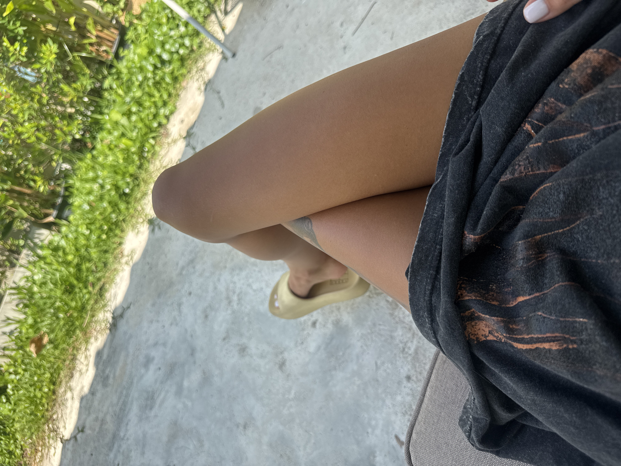 The image, taken from a high angle, shows a person's legs and part of their torso. The person is wearing a dark-colored, possibly tie-dyed, garment that covers their lap. Their legs are bare, and they have a tan. On one foot, they are wearing a yellow or beige shoe. A small tattoo is visible on the lower leg. The background consists of a concrete surface and some greenery along the side. The setting is likely outdoors. The location given is Choeng Thale, Thailand, but there is no specific visual information in the image to confirm that location beyond the vegetation potentially typical of the area.