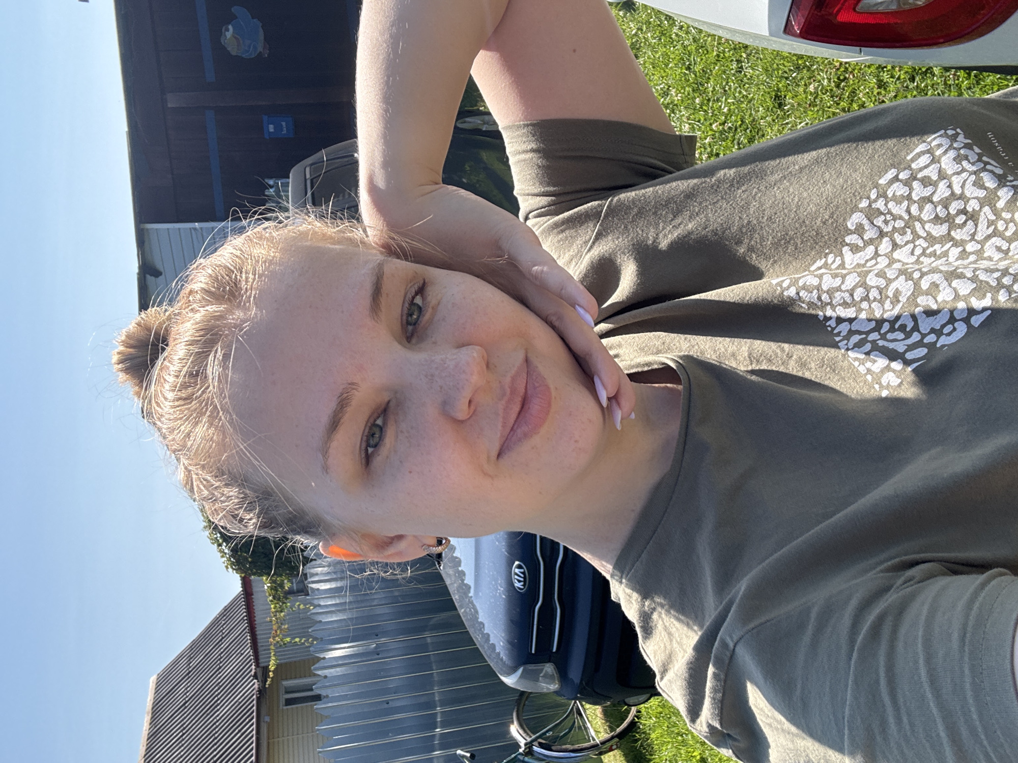 A young woman with blonde hair pulled into a bun and light skin with freckles is taking a selfie outdoors. She is wearing a green t-shirt with a white, abstract pattern on it. Her right arm is bent, and her hand is resting on her cheek, supporting her head. She is smiling slightly and looking at the camera.
In the background, a dark blue Kia Soul car is parked, with a bicycle leaning against a metal fence. Behind the fence, a yellow house with a grey corrugated roof is visible, and further back, a dark wooden structure with a blue bird painted on it. The ground is covered in green grass, and the sky is clear blue, indicating a sunny day. The setting appears to be a rural area, possibly a village like Дунилово, Russia, given the presence of a house, a fence, and outdoor parking.